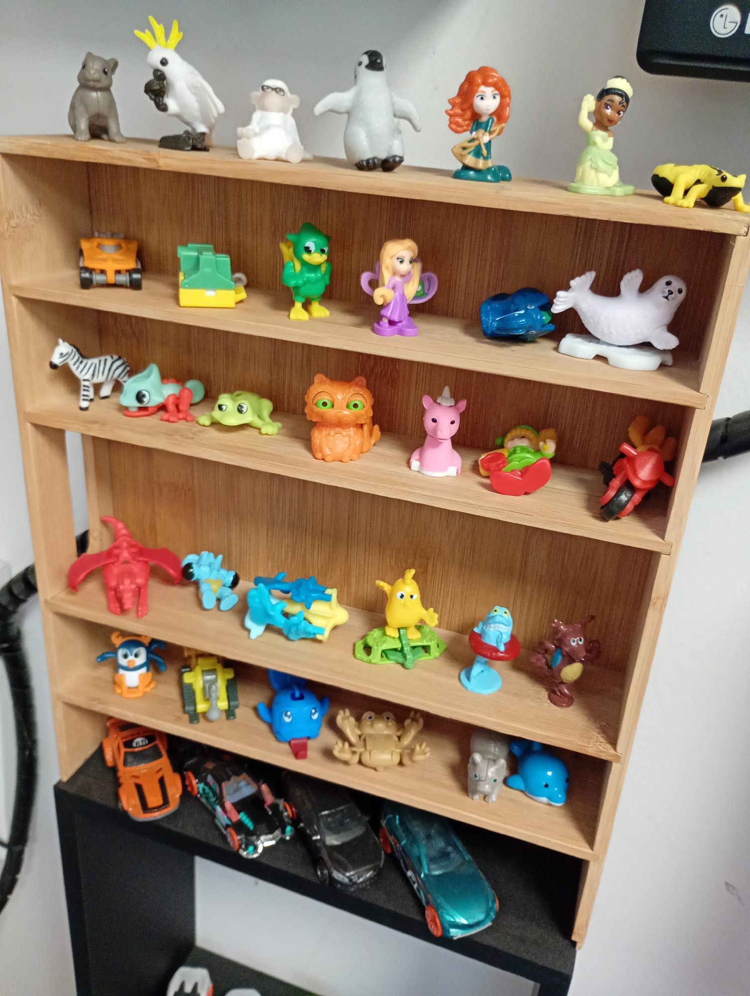 A detailed view captures a collection of small plastic toy figurines neatly arranged on a multi-tiered wooden display shelf. The shelf, made of light-colored wood with visible grain, features five distinct levels, with additional toys on a black shelf or base below.
The figurines are diverse, primarily consisting of animal figures, fantasy creatures, and a few human-like characters or dolls, alongside several miniature vehicles. On the topmost shelf, from left to right, are a grey bear-like animal, a white cockatoo, a white monkey-like figure with glasses, a white penguin, a redhead princess doll (resembling Merida), a dark-skinned princess doll (resembling Tiana), and two yellow frog-like creatures.
The subsequent shelves showcase an array of items: an orange construction vehicle, a green and yellow construction toy, a green bird-like creature, a purple fairy-like doll, a blue car, and a white seal on the second level. The third shelf holds a zebra, a green lizard, a green frog, an orange owl-like creature, a pink unicorn, a baby in a red bath, and a red motorcycle. The fourth shelf displays a red dragon, a blue creature/robot, a blue and yellow creature, a yellow bird, a blue frog in a red float, and a brown creature. The lowest wooden shelf features a blue and orange penguin, a yellow excavator, a blue robot, a tan frog, a grey rhinoceros, and a blue dolphin. On the black base below, four miniature cars are lined up: an orange car, a black car with colorful details, another black car, and a teal/green car.
The background is a plain, light-colored wall, and a black cable or hose is partially visible on both sides of the display. No activity is taking place; the toys are static and meticulously presented. The lighting is even, indicating an indoor setting. A white "LG" logo is partially visible on a black object in the top right corner of the image.
