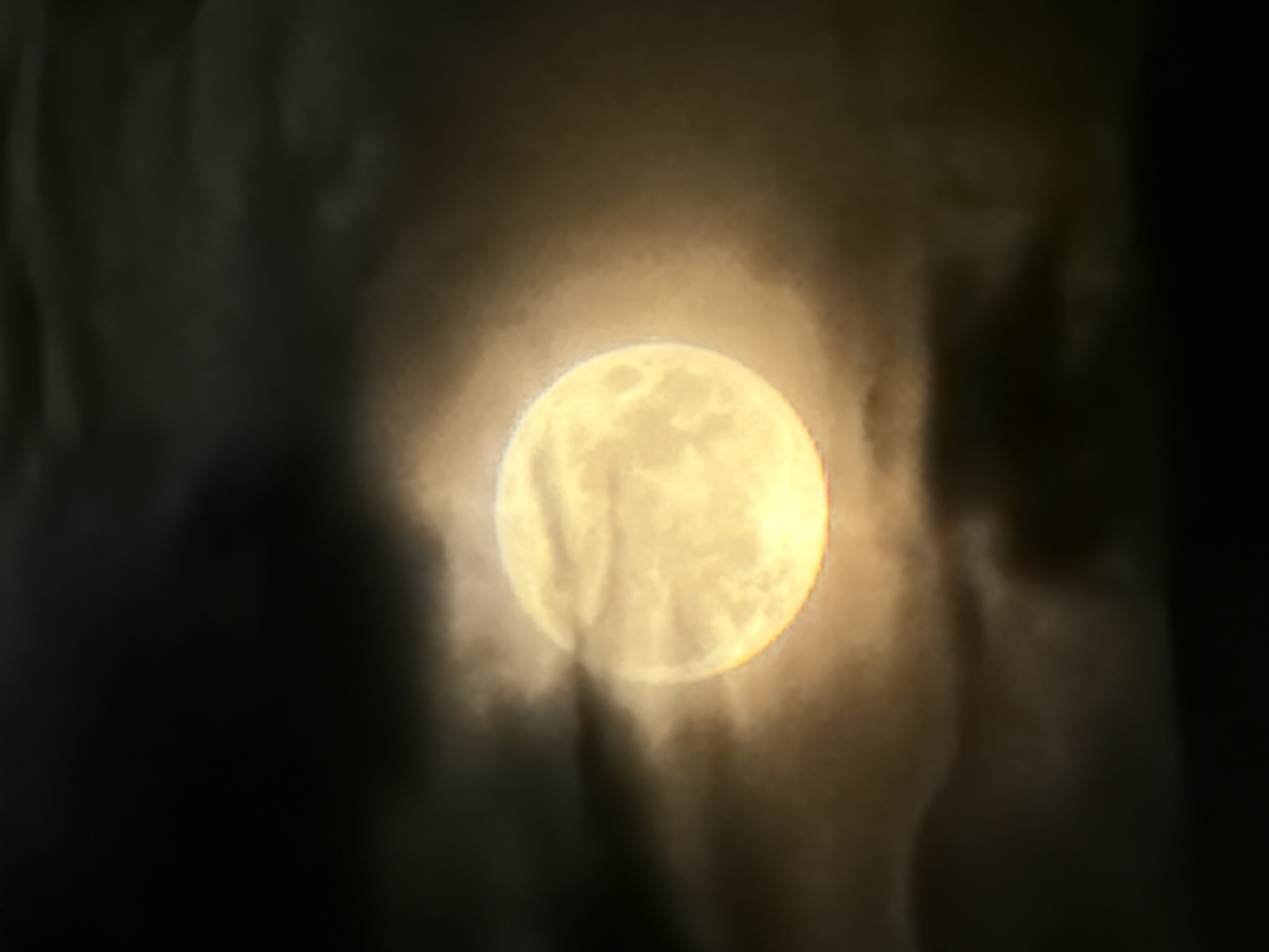 A full moon is visible through a layer of dark, diffuse clouds. The moon is bright yellow, with visible surface texture. The surrounding clouds are dark gray and appear to obscure the moon's edges. There is no information to verify the location of the city of Chaikovsky, Russia.