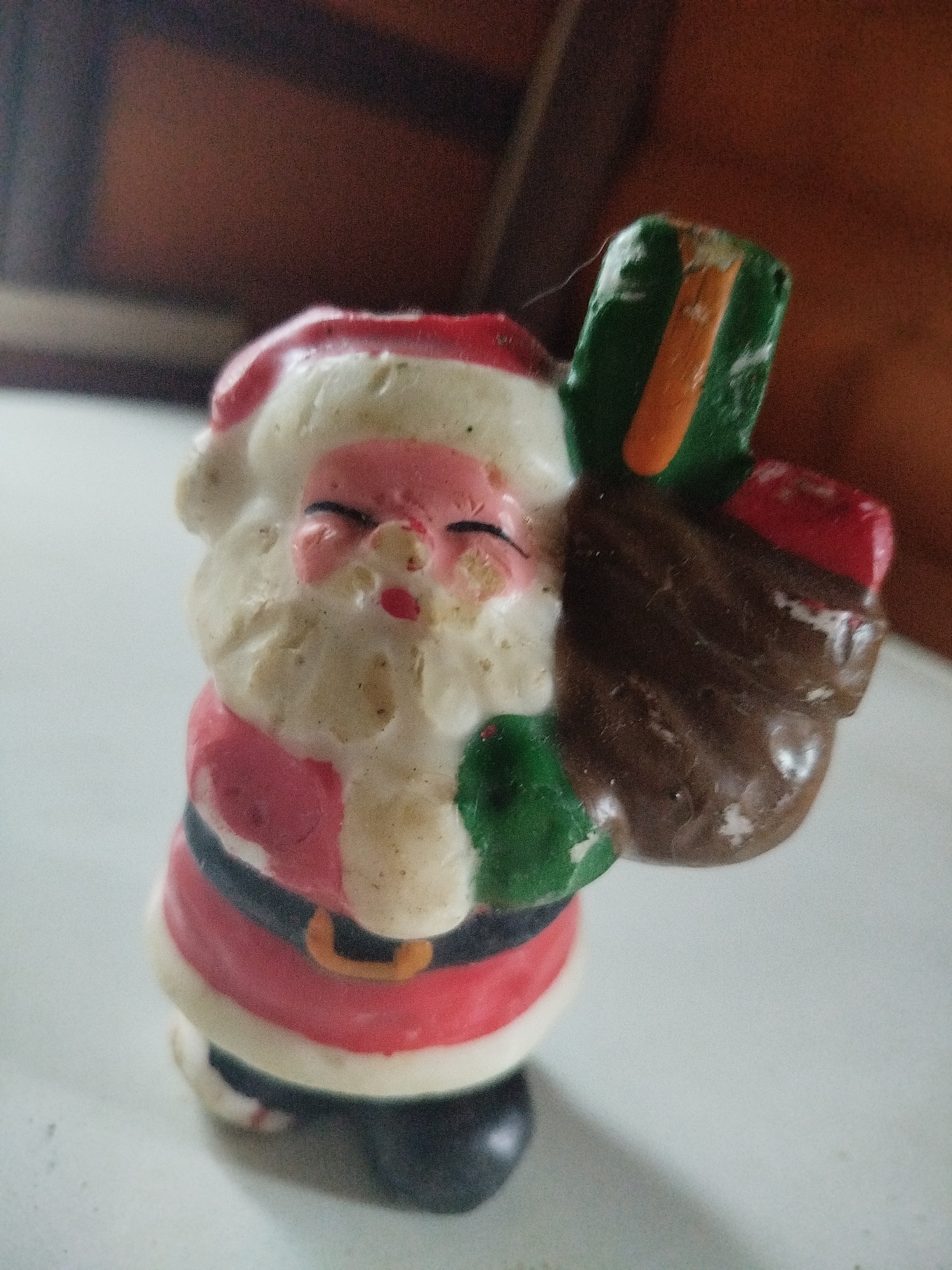 An old, slightly deteriorated Santa Claus figurine stands on a light-colored surface. The figurine is made of painted plaster or a similar material, with some discoloration and wear visible. Santa has a red hat with a white brim, a pink face with closed eyes, and a white beard. He is wearing a red suit with a white trim and a black belt with a gold buckle. On his back, he carries a brown sack filled with presents, including a green gift with an orange ribbon.
There is no information in the image that indicates the location is Боровково, Russia.