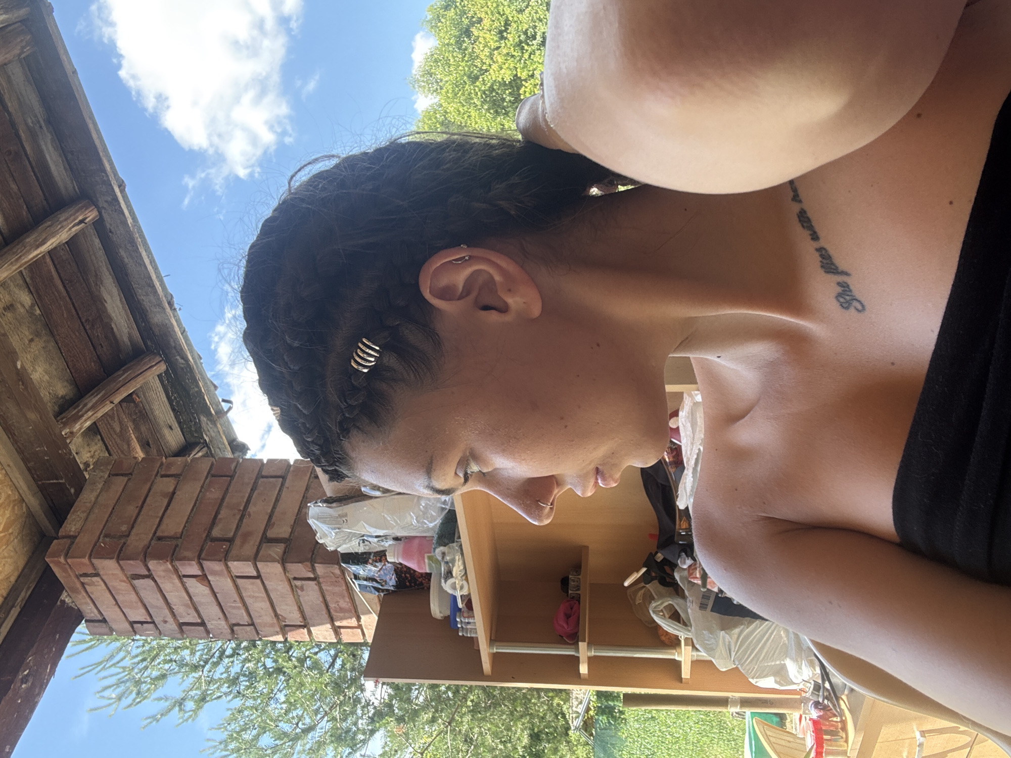 In Belopoptsi, Bulgaria, a woman with braided dark hair, adorned with several silver rings, sits outdoors under a wooden roof. She is wearing a black strapless top and has a tattoo on her collarbone. The background reveals a brick pillar, shelves filled with various items, and lush green trees under a bright blue sky with white clouds. The sunlight casts a warm glow on the scene.
