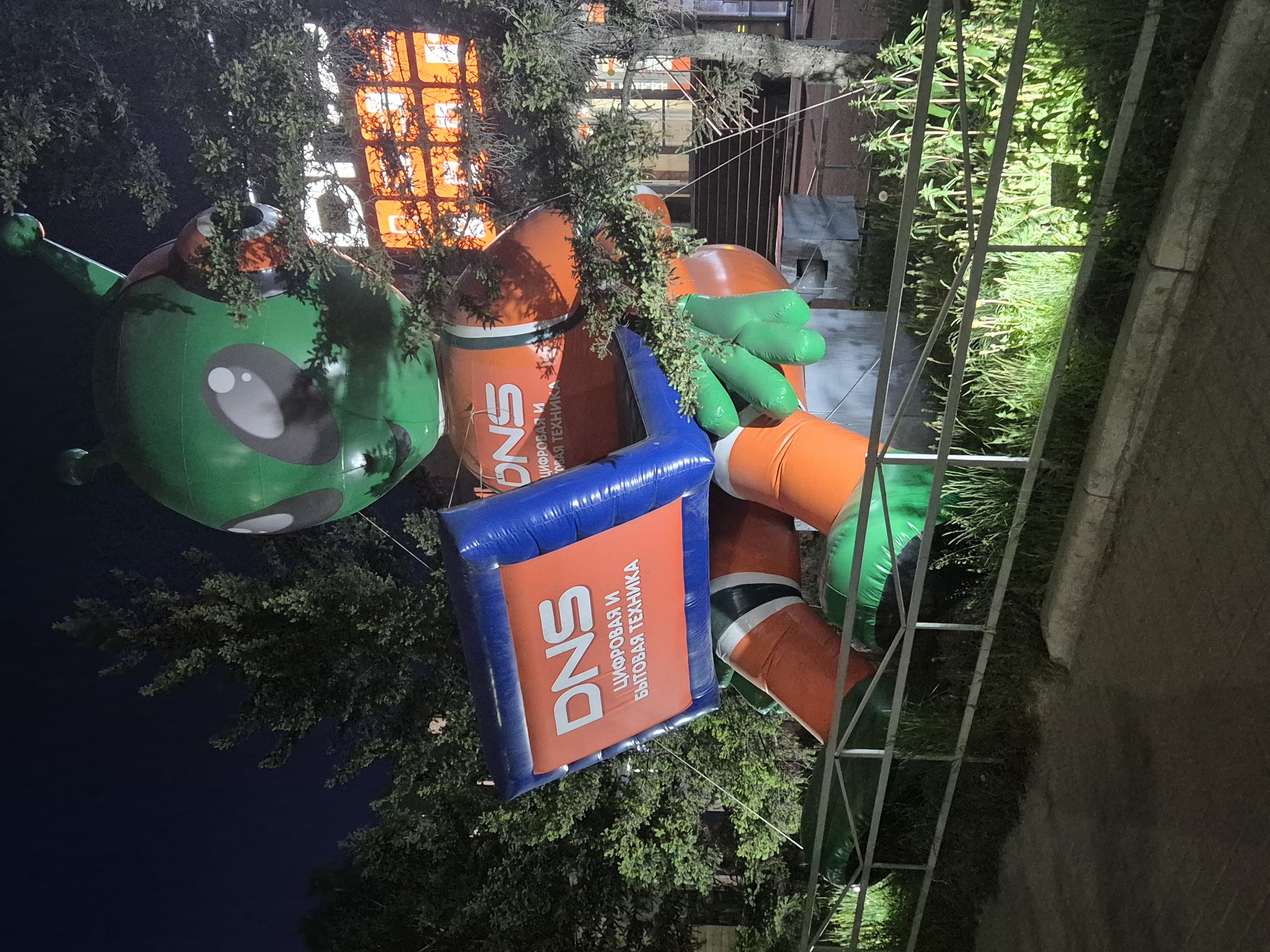The image captures an outdoor scene at night, featuring a cluster of inflatable advertising figures, including a green alien balloon with a large eye, and cylindrical orange inflatables adorned with the DNS logo (a Russian electronics store). A blue inflatable sign, also with the DNS logo, hangs slightly askew, partially obscured by foliage. The inflatables are positioned amidst trees. In the background, a building displays lit windows. Adjacent to the inflatables is a metal fence or railing, with grass and foliage growing nearby. The location appears to be in the city of Unknown, Unknown.