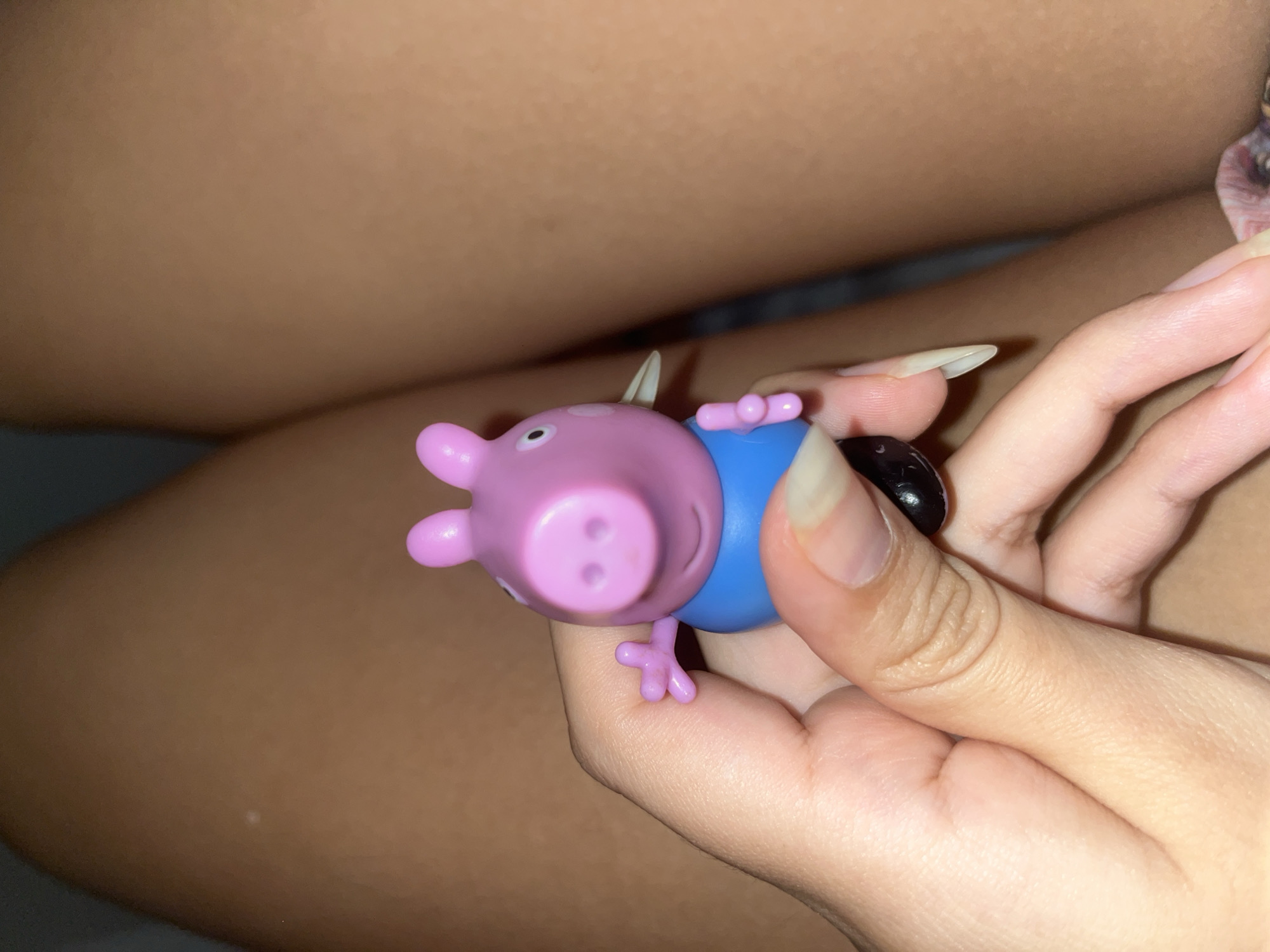 A close-up shot features a person's hand holding a Peppa Pig toy. The toy is predominantly pink with a blue shirt, and its face is clearly visible. The person's skin tone is fair, and their nails are long and manicured with what appears to be a light-colored polish. The background is blurred, showing a tan surface, possibly human skin, and a hint of fabric with a floral pattern. The image is not taken in the city of San Luis, Peru, and there is no indication of the location in Peru.