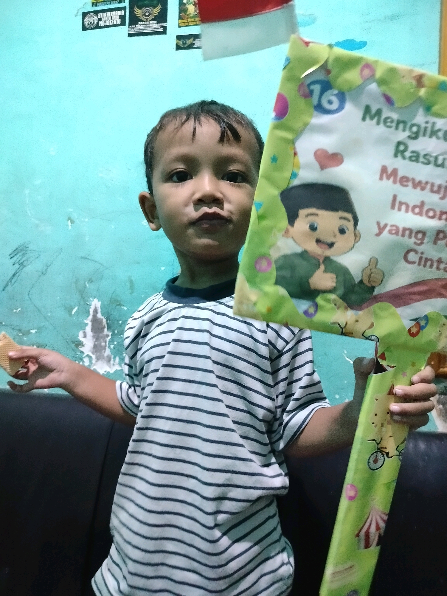 A young boy stands in front of a light blue wall in Beloh, Indonesia. He is wearing a black and white striped t-shirt and holding a colorful poster with a cartoon of a boy giving a thumbs-up. The poster has text in Indonesian, including "Mengikuti Rasulu" and "Mewujudkan Indonesia yang Pancasila". The wall behind him is decorated with several stickers or posters. To the left of the boy, a piece of a wafer is visible in his hand. The lighting suggests the photo was taken indoors.