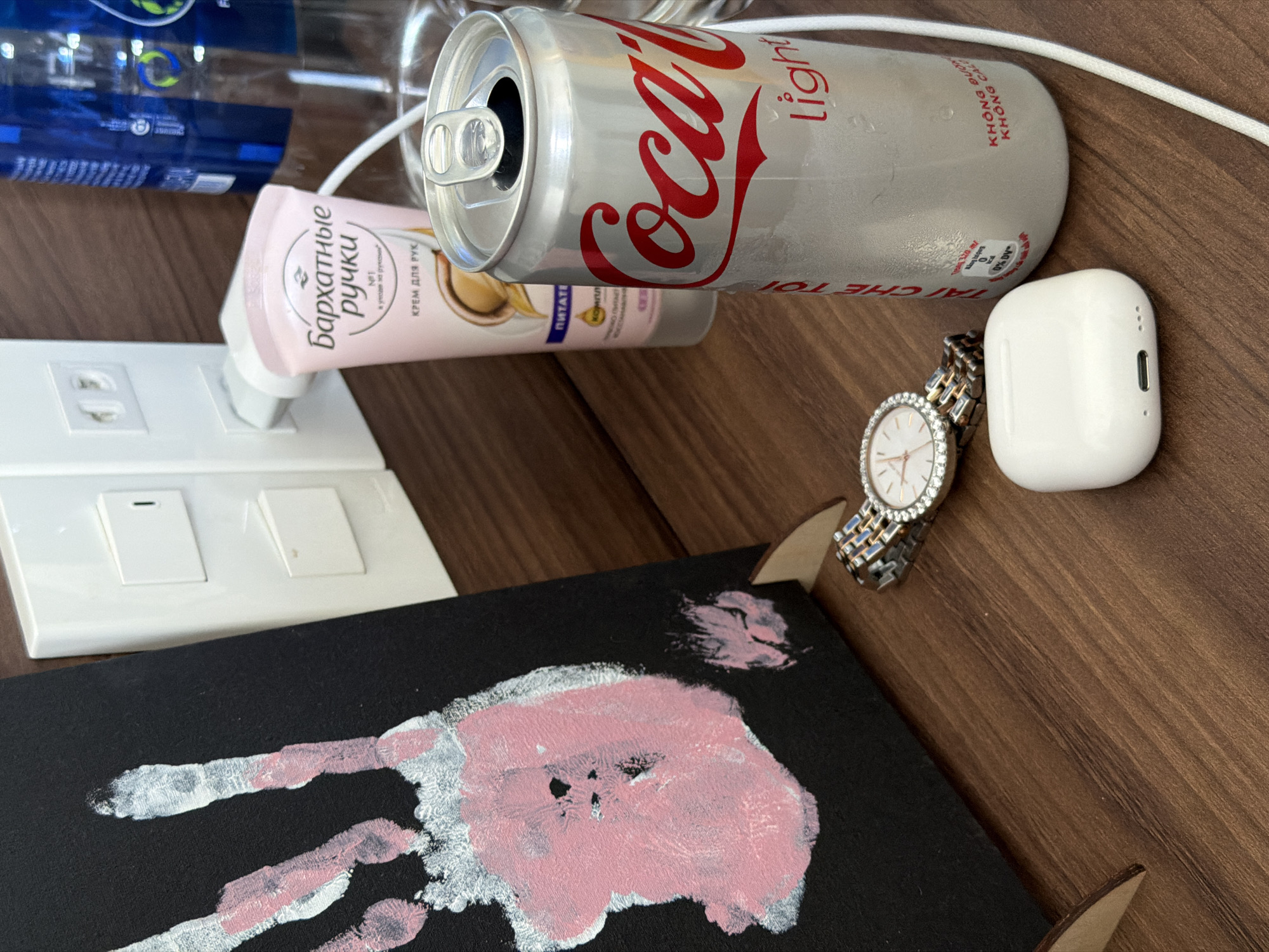 The image shows a table with several items on it. A half-empty bottle of water, a can of Coca-Cola Light, and a tube of Russian hand cream are placed near a white light switch plate. Next to these items are a watch with a silver and gold band, a white AirPods case, and a piece of black paper with a child's handprint in pink and silver paint. The surface of the table appears to be wood. Based on the Vietnamese text on the Coca-Cola can, it is possible this image was taken in Vietnam. Without further evidence, it is impossible to say for certain this image was taken in Đà Nẵng, Vietnam.