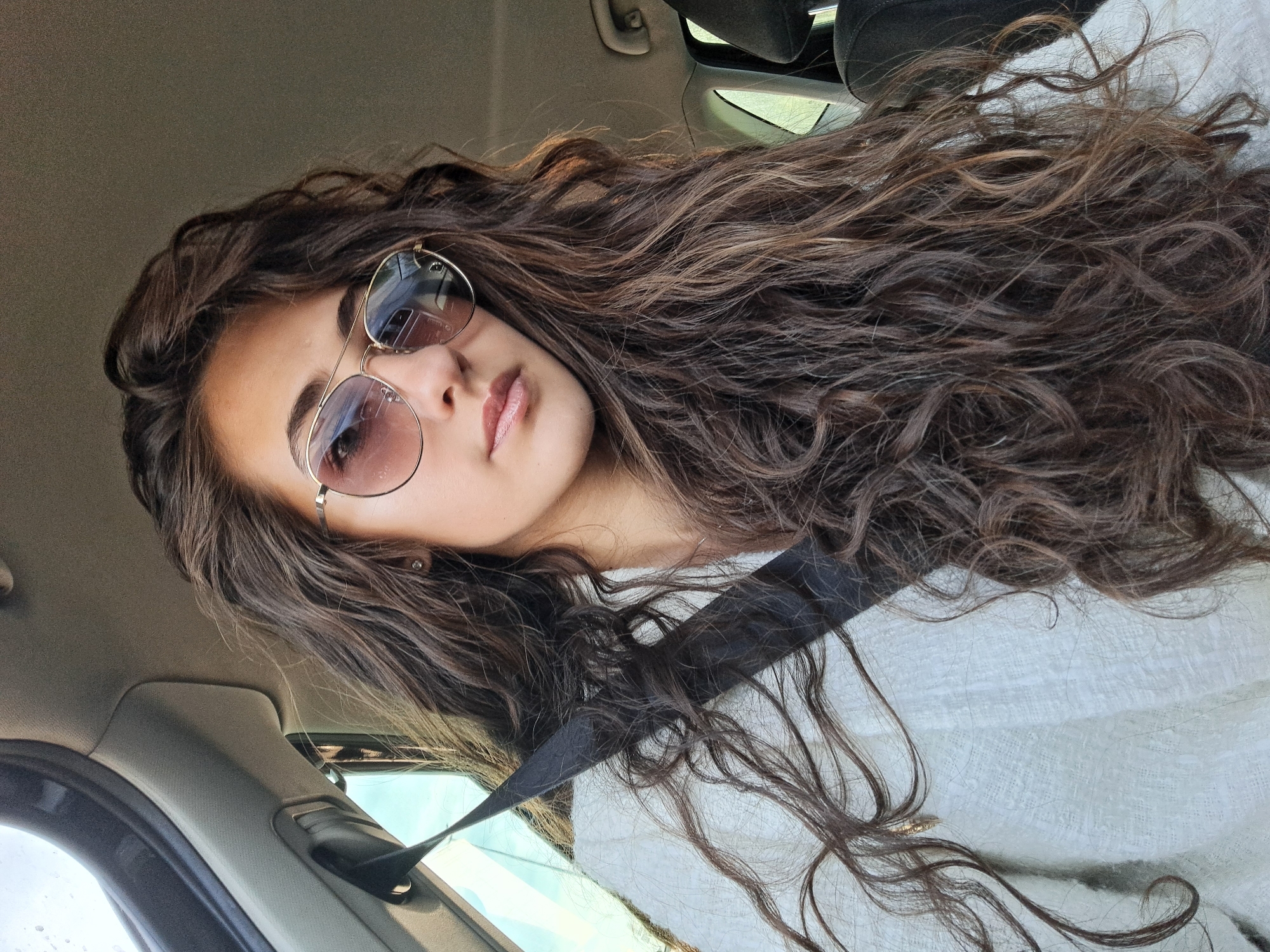 A woman with long, curly brown hair is in the driver's seat of a car, wearing a white sweater and sunglasses. She has a seatbelt on. The location is not discernible as Krivina, Bulgaria from this image alone. The focus is primarily on the woman and the interior of the vehicle.