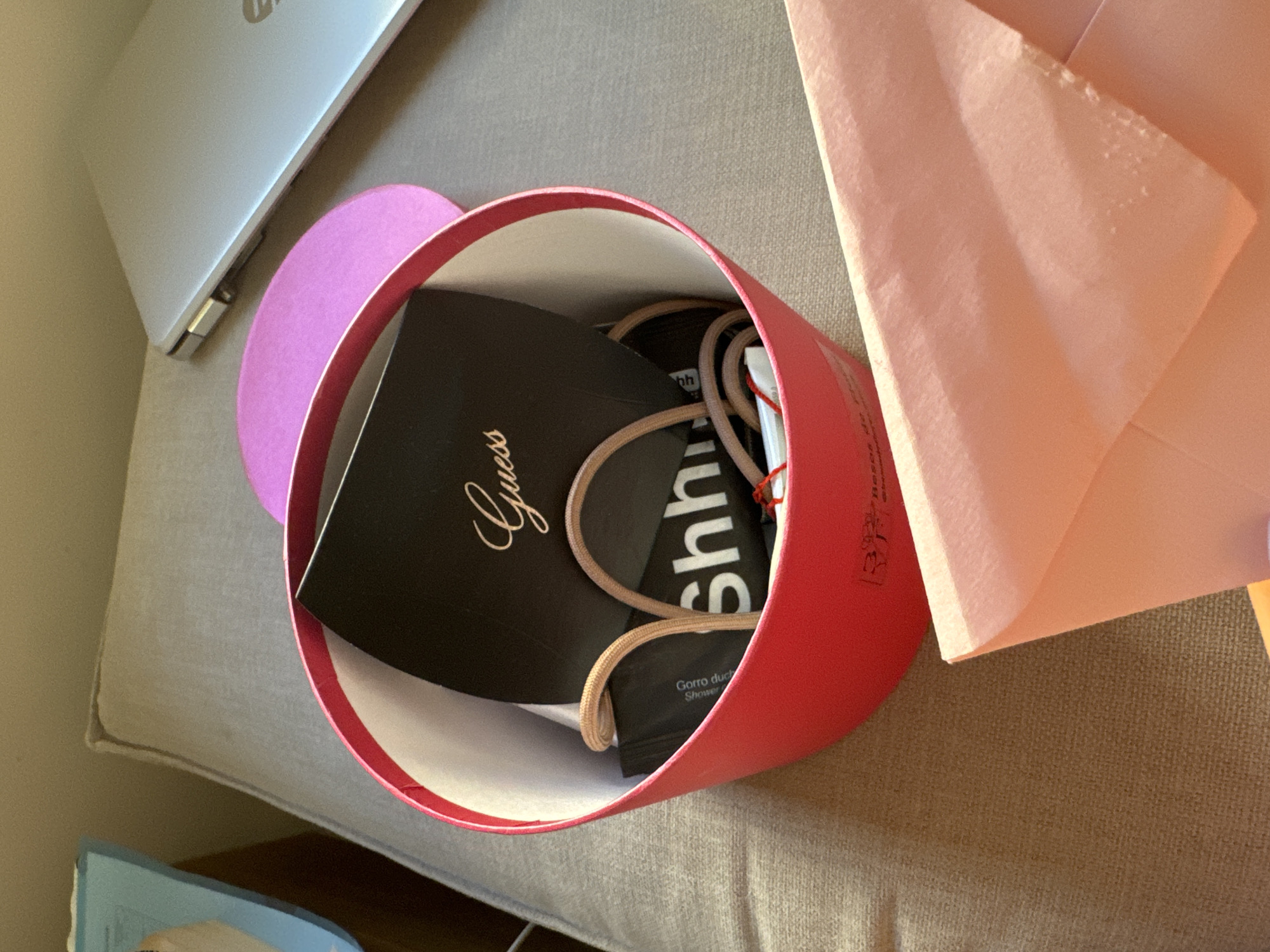 The image shows a red cylindrical gift box filled with various items, including a black card with the "Guess" logo and "Shhh" printed on it, and two tan drawstrings. A pink circular lid rests next to the box. A portion of a silver laptop is visible in the background, along with a light blue object on the left. In the foreground, a pale pink envelope is partially visible. The scene is set on a textured, neutral-colored surface, likely a sofa or upholstered furniture. The lighting suggests an indoor environment. There is no indication of the city of Murcia, Spain, or any specific location within it. The image focuses on the contents of the gift box and surrounding objects.