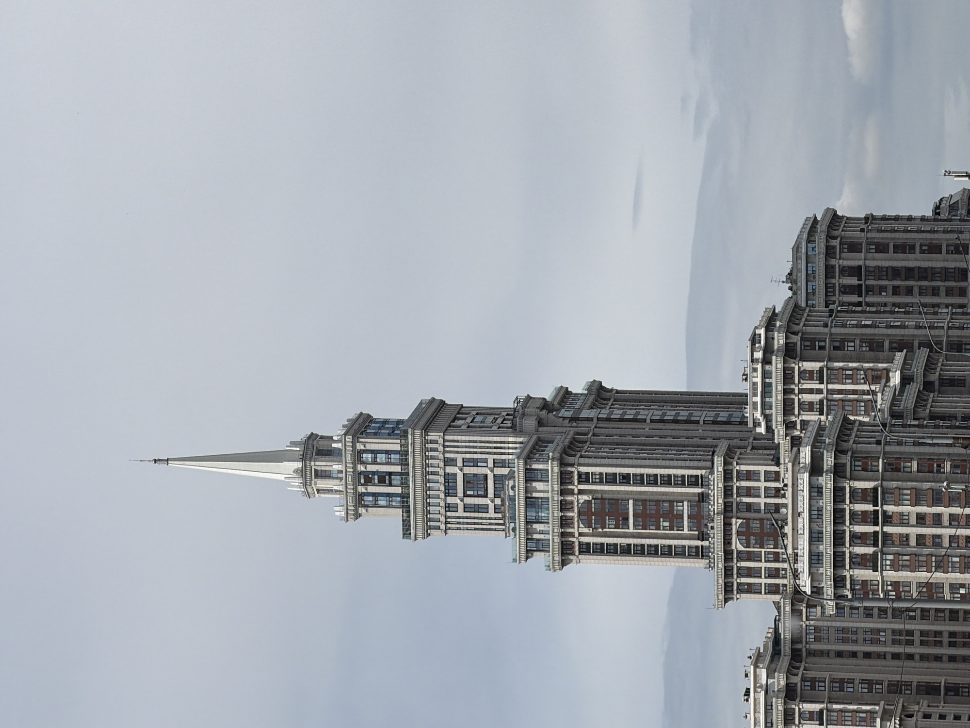 A tall, ornate skyscraper with a spire dominates the center of the frame, its facade a mix of grey stone and dark-tinted windows. The skyscraper is flanked by other buildings of similar architectural style, though shorter. The sky above is overcast with light grey clouds. The scene appears to be a cityscape.