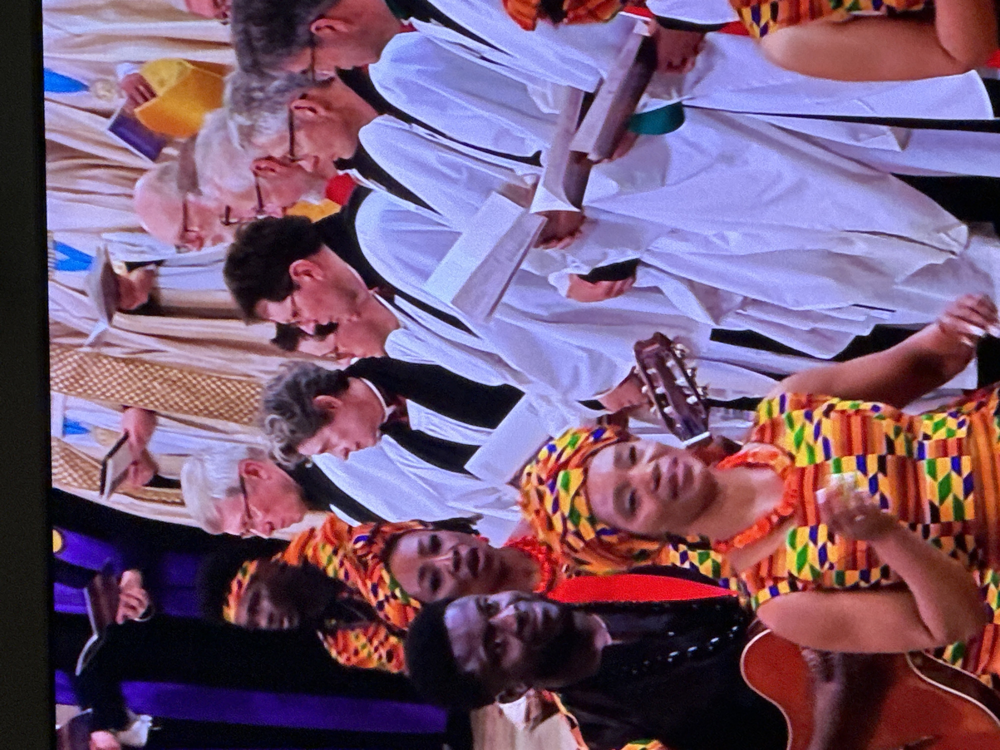 The image depicts an indoor scene within a formal religious building in London, United Kingdom. Multiple individuals are arranged in rows, participating in what appears to be a service or performance.
In the foreground, a woman of African descent wears a vibrant Kente-patterned sleeveless dress and a matching headwrap, complemented by an orange beaded necklace. She smiles slightly while holding a small, light-colored object in her right hand. To her left and slightly behind, a man of African descent, wearing a black top, holds an acoustic guitar. He smiles and looks towards the viewer. Behind him, another woman of African descent, wearing a Kente-patterned headwrap and a red top with an orange beaded necklace, has her mouth slightly open. Further back, a fourth individual of African descent wears a Kente-patterned headband and shirt.
Behind and interspersed among these individuals, numerous other participants are visible. These are primarily older white males, uniformly dressed in white ecclesiastical robes (albs), black clerical stoles, and collars. Many hold open books, appearing to be hymn books or service programs. Some display additional liturgical or academic attire, including academic hoods; one individual wears a prominent gold and purple hood. The individuals in robes stand formally, generally facing forward.
The environment is brightly lit, indicating a large, public interior space within a significant institution.