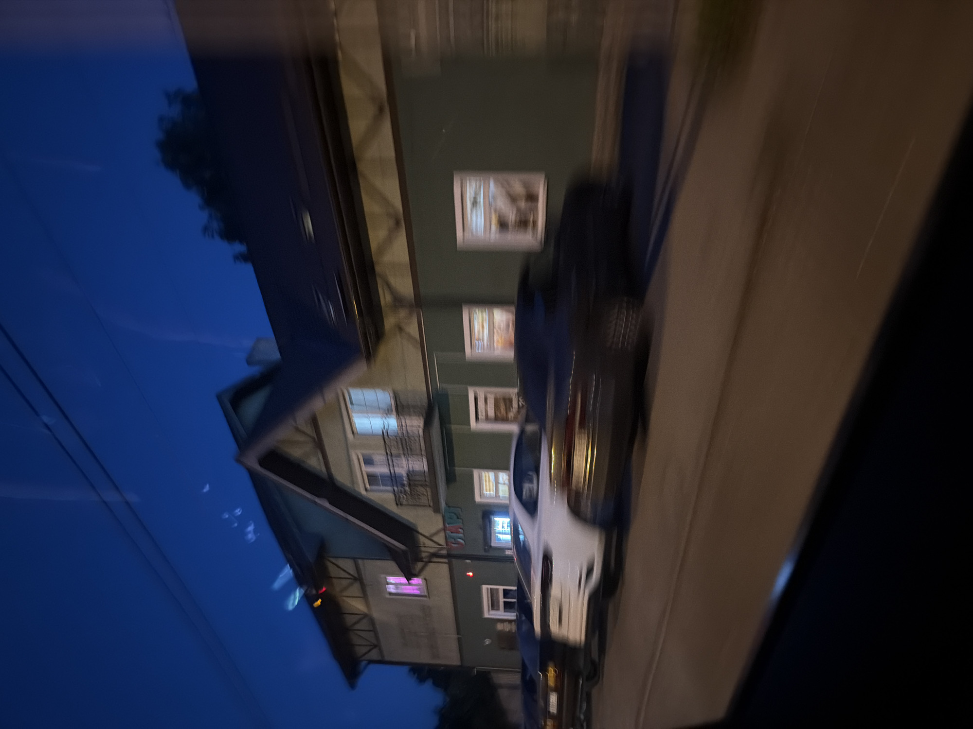The image captures a street scene at dusk, likely in Kaliningrad, Russia, given the presence of Cyrillic text. A two-story building with a green facade and a dark roof is prominent. The building features several windows, some of which are illuminated, and a balcony on the upper floor. A sign with the Cyrillic letters "CLAP" is visible on the building's facade. In front of the building, several cars are parked or driving along the street, their forms blurred due to motion. The sky is a deep blue, suggesting it is either early evening or late twilight. The overall impression is a quiet street in a European city at the end of the day.