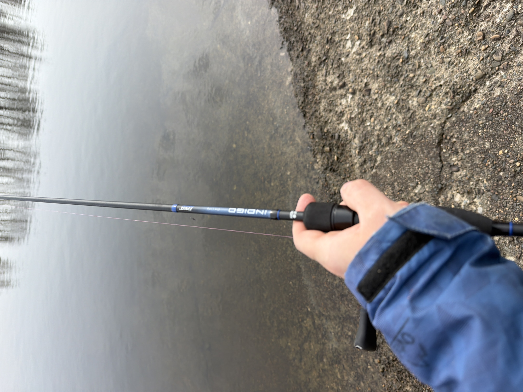 A first-person view shows an individual holding a fishing rod, standing near the edge of a body of water. The fishing rod is dark, with blue accents and the word "INDIGO" printed on its side. A thin, pink fishing line extends from the rod. The individual is wearing a blue jacket with a velcro closure on the sleeve.
The environment consists of water on one side and what appears to be a rocky or gravelly bank on the other. The water surface is somewhat reflective, and some trees are visible in the distance.
The location is Chaikovsky, Russia.