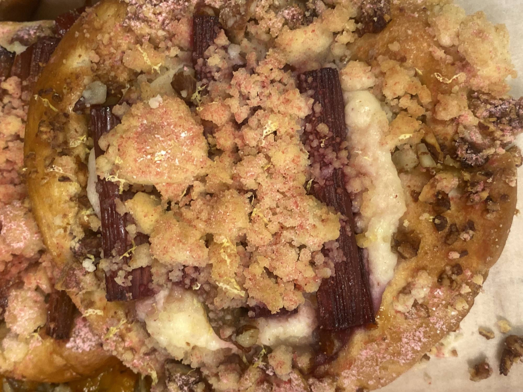 This is a close-up, overhead shot of a baked good, likely a pastry or tart, placed on a white surface. The pastry has a golden-brown crust, with visible crumbles of topping scattered across its surface. The topping appears to be a mixture of what looks like streusel, possibly made with flour, sugar, and butter, and it's flecked with pink and yellow particles, suggesting the presence of fruit or coloring.
Dominating the center of the pastry are dark red, elongated pieces, which are consistent with rhubarb stalks. These are interspersed with dollops of a white, creamy filling. Scattered amongst the toppings and crust are small, dark brown pieces, which could be nuts, such as walnuts or pecans.
The overall impression is of a rustic, homemade dessert. There are no people visible, and the setting is a simple white surface, providing no specific location cues beyond suggesting it might be a kitchen counter or serving dish. The lighting is even and appears to be artificial, offering no clues about the time of day or weather. The emotions evoked are generally positive, associated with the enjoyment of a sweet treat. There is no visible text in the image.