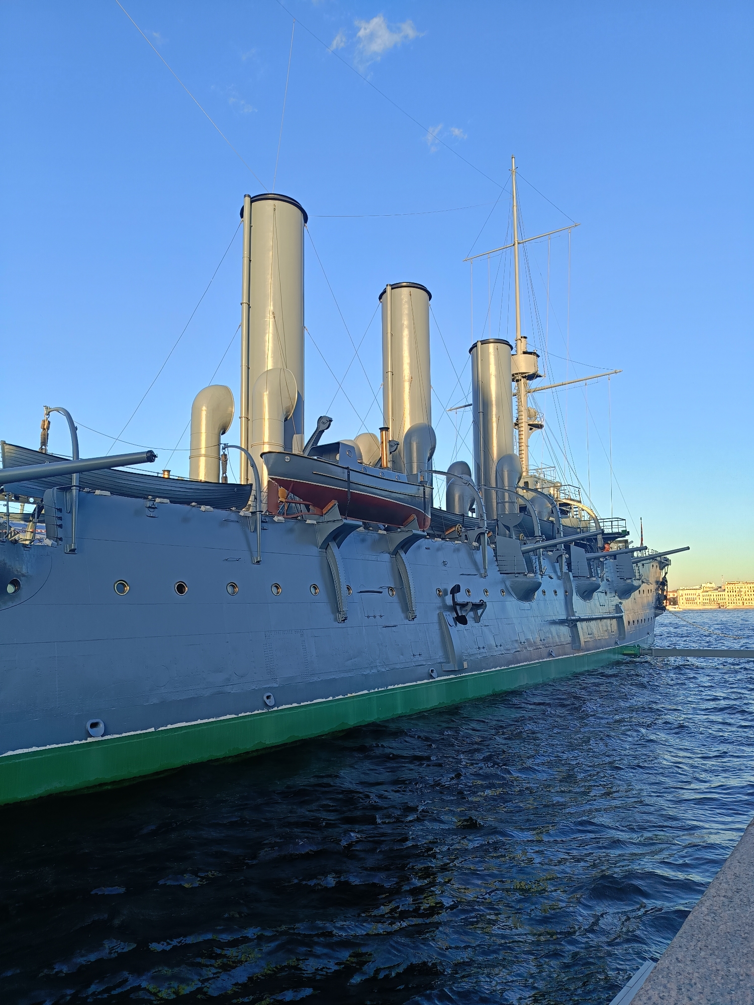 This is a photograph of the historic cruiser Aurora, moored in the Petrovskaya Embankment of the Neva River in Saint Petersburg, Russia. The ship is painted a muted gray with a vibrant green waterline. Four large grey smokestacks dominate the upper deck, with intricate rigging and masts rising behind them. A small life raft is suspended from davits on the starboard side, alongside other deck equipment. The ship's hull features a row of circular portholes and several large gun turrets. The calm, dark blue water of the Neva River laps against the hull and a stone embankment is visible in the foreground. In the background, across the river, a cluster of light-colored buildings with a characteristic Saint Petersburg architectural style can be seen under a clear, light blue sky with a few wispy clouds. The lighting suggests it is late afternoon or early evening. There are no people visible in the frame.