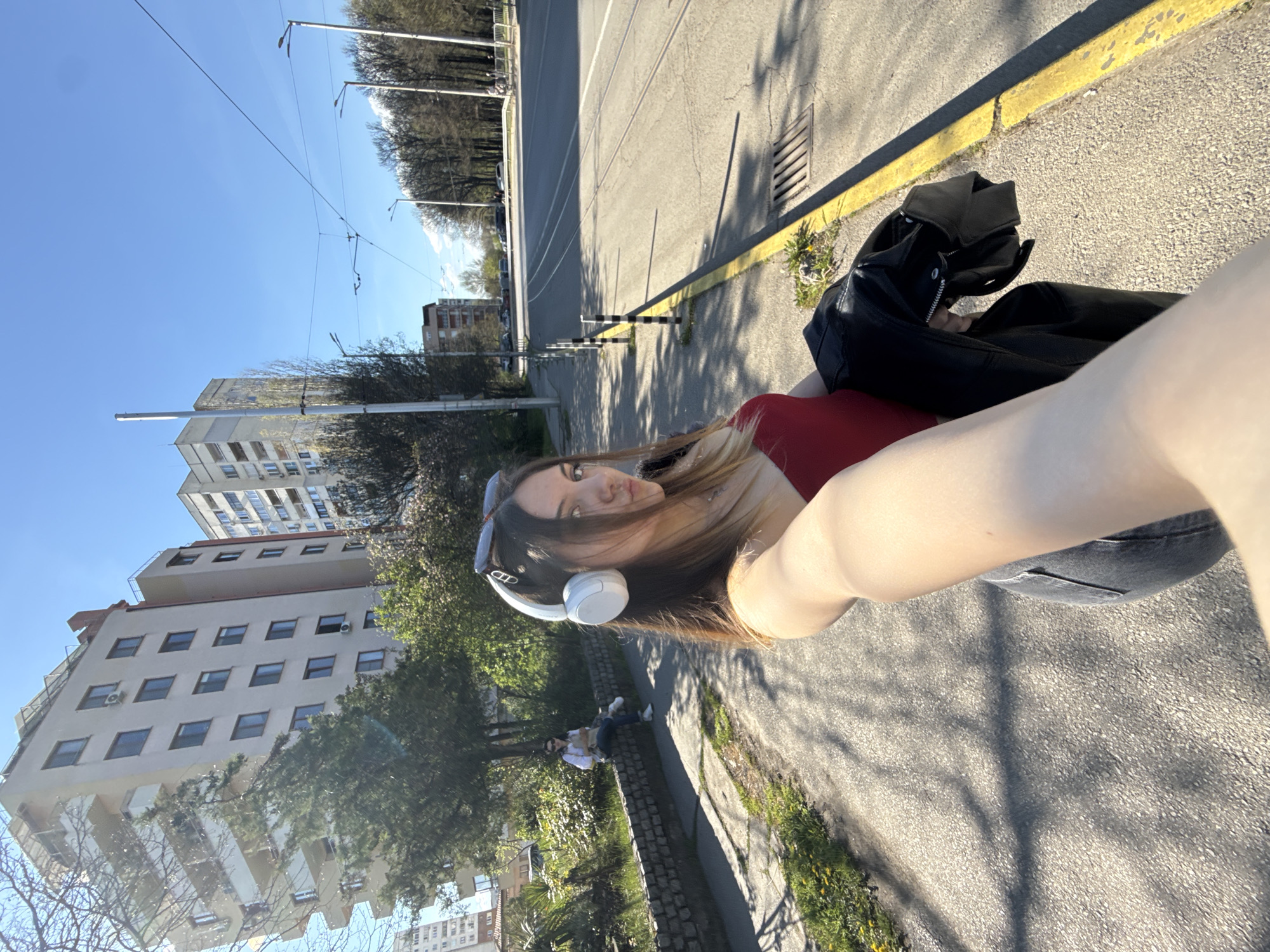 The image is a selfie taken by a young woman. She is holding a black leather jacket. She is wearing white headphones and a red t-shirt. She has sunglasses on her head. In the background, there is a street with tram lines, buildings, and trees. Two people are sitting on a low wall. The location is likely Sofia, Bulgaria, given the architecture.