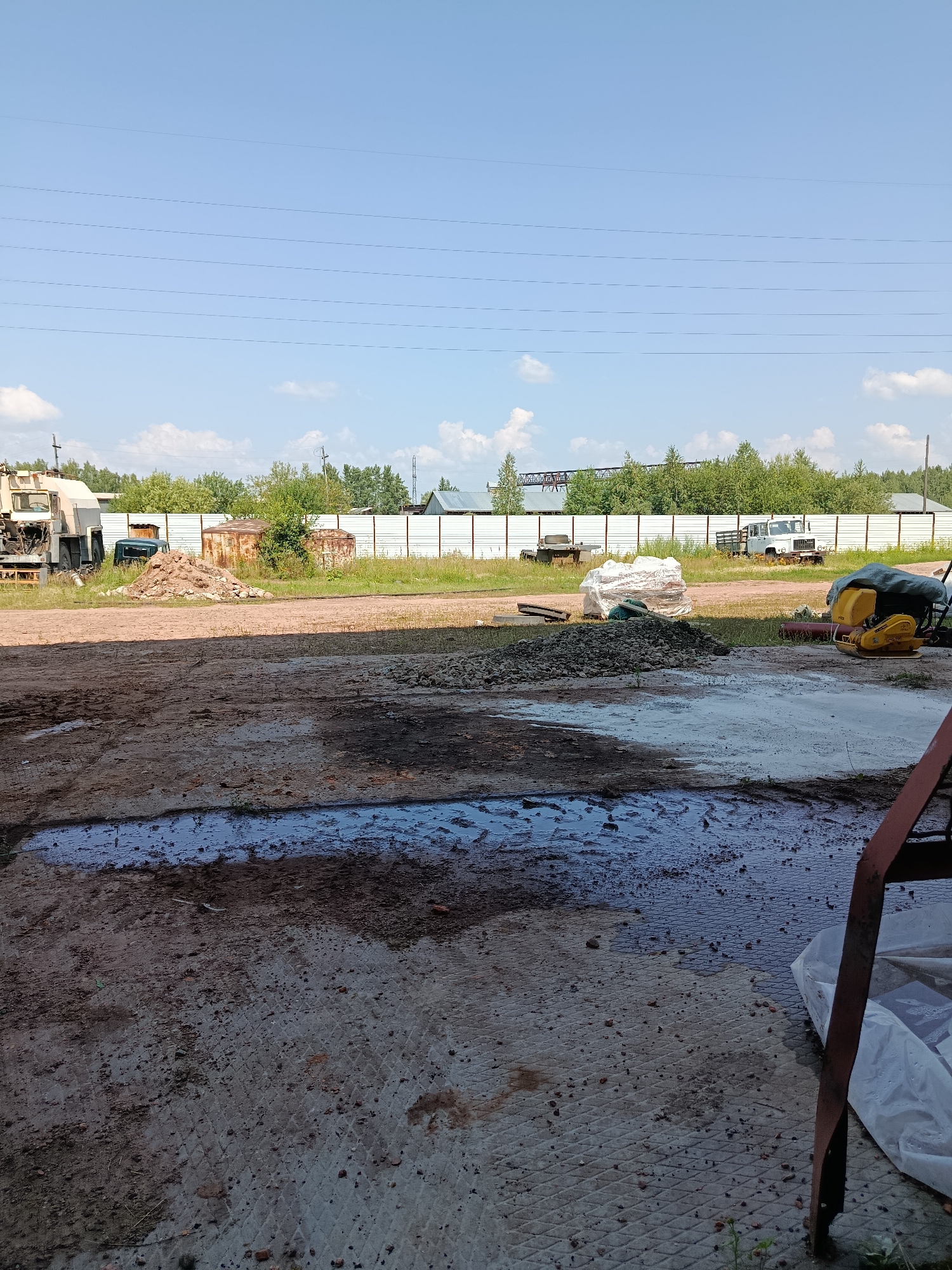 The image depicts an outdoor industrial or construction area in Borodino, Russia. In the foreground, a dirt and concrete ground surface is visible with puddles of water and scattered debris. Further back, there is a pile of gravel and a yellow compaction machine covered with a tarp. Several vehicles, including a truck and what appears to be a small utility vehicle, are parked in the mid-ground. A white fence runs along the edge of the property, with a line of trees and industrial structures visible beyond it. The sky is clear with some scattered clouds. The overall scene suggests a working environment with building materials and equipment.