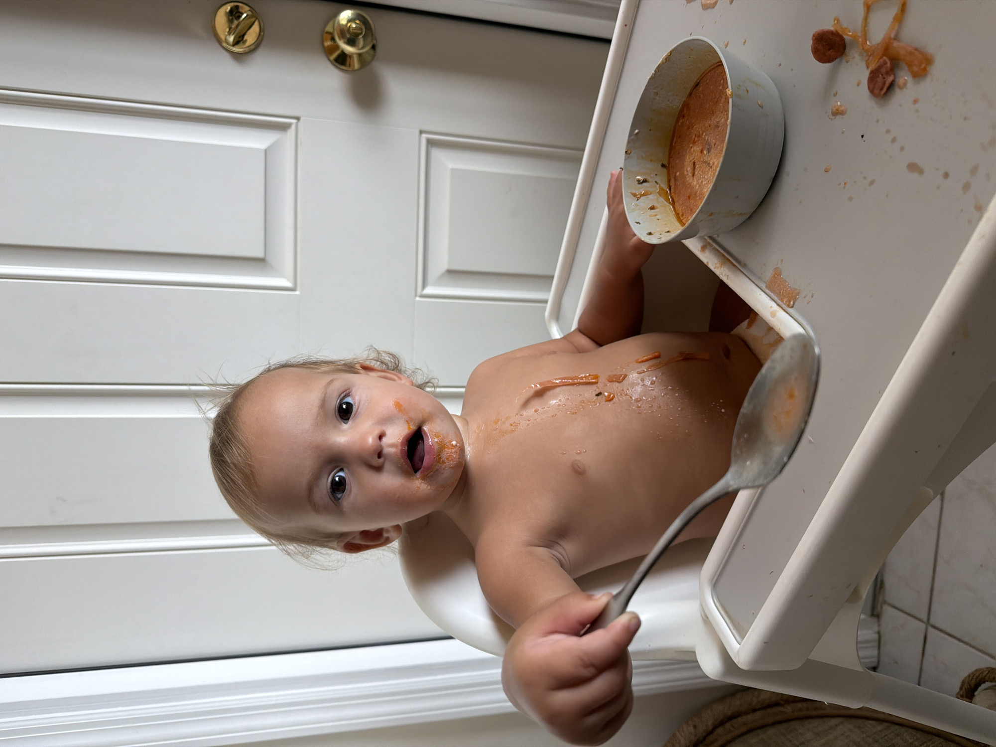 A baby sits in a highchair with food spilled on their chest and face, holding a spoon. A bowl of food sits on the highchair tray along with spilled food. A white door with gold hardware is visible in the background. The location is not specified in the image and cannot be identified.