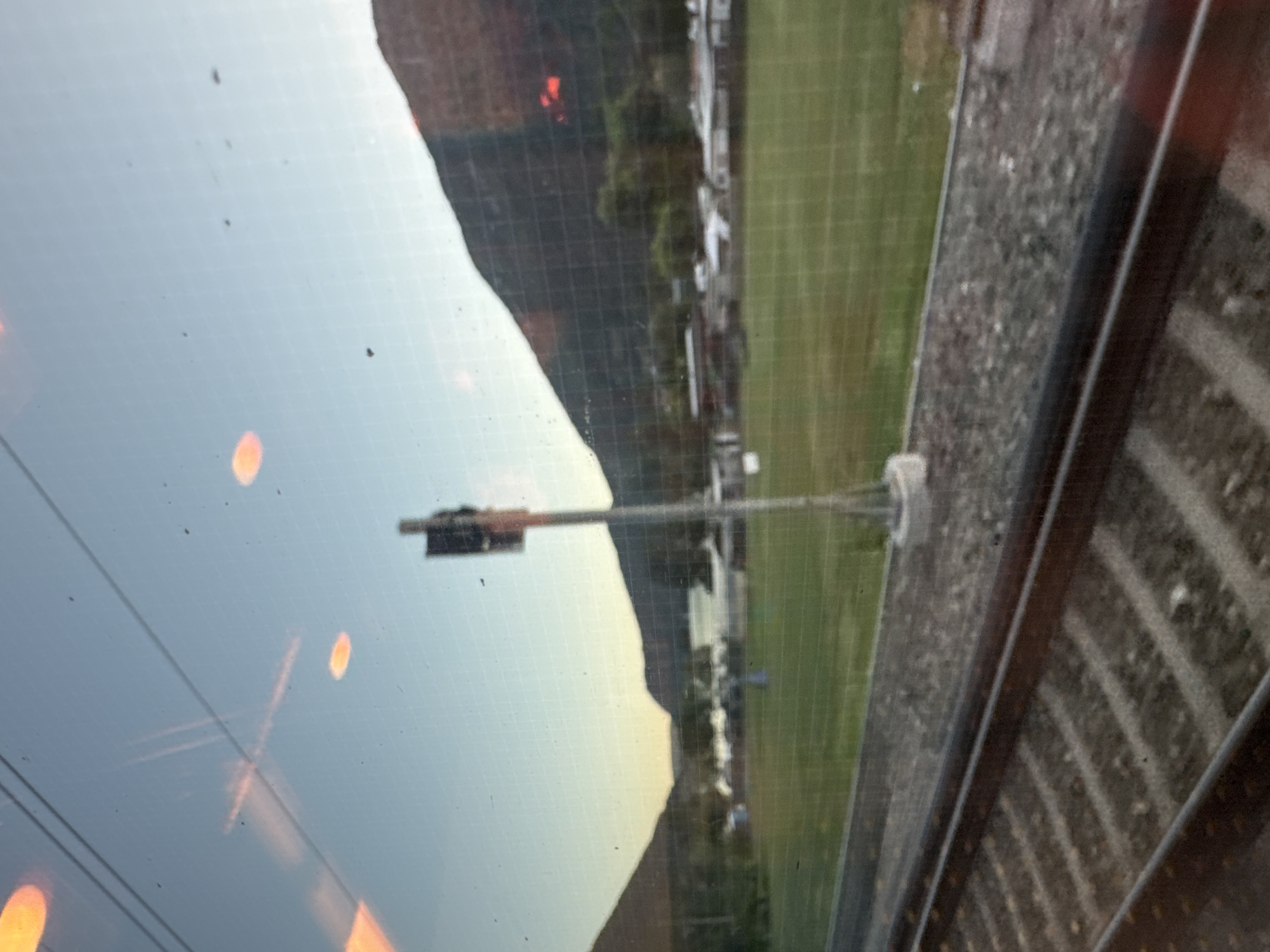 The image depicts a view from a train, looking out at a landscape with a body of water in the distance, framed by hills or mountains. The foreground shows train tracks and ballast. Beyond the tracks is a grassy field, and further back, there appears to be a small settlement or campground with several structures, possibly including tents and buildings. A tall pole with a signal or light is positioned in the field. The sky is clear and shows a gradient of blue and lighter tones, suggesting either dawn or dusk. There are reflections on the window of the train, including orange circular shapes and linear patterns. The location is identified as the city of Val Mara, Switzerland.