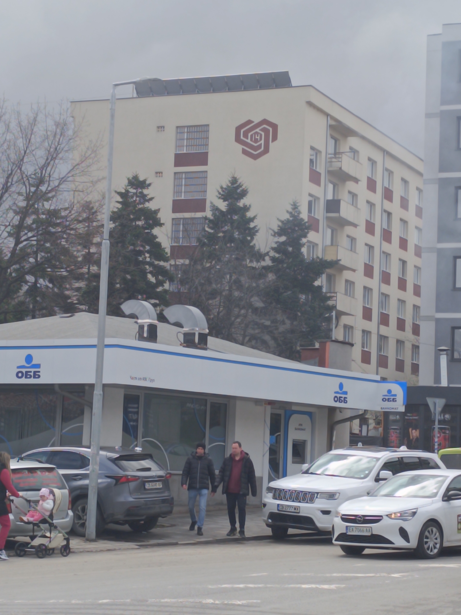 The image depicts a street scene in Sofia, Bulgaria. The primary focus is on an OBB (United Bulgarian Bank) branch, characterized by its white and blue branding. Several vehicles are parked in front of the bank, including a gray SUV, a white Jeep, and a white Opel Corsa. Pedestrians are present: a woman pushing a stroller, and two men walking side-by-side.
In the background is a beige building adorned with a symbol resembling interconnected stylized hearts with the number "14" inside. The building has multiple floors with red-framed windows. The weather is overcast.