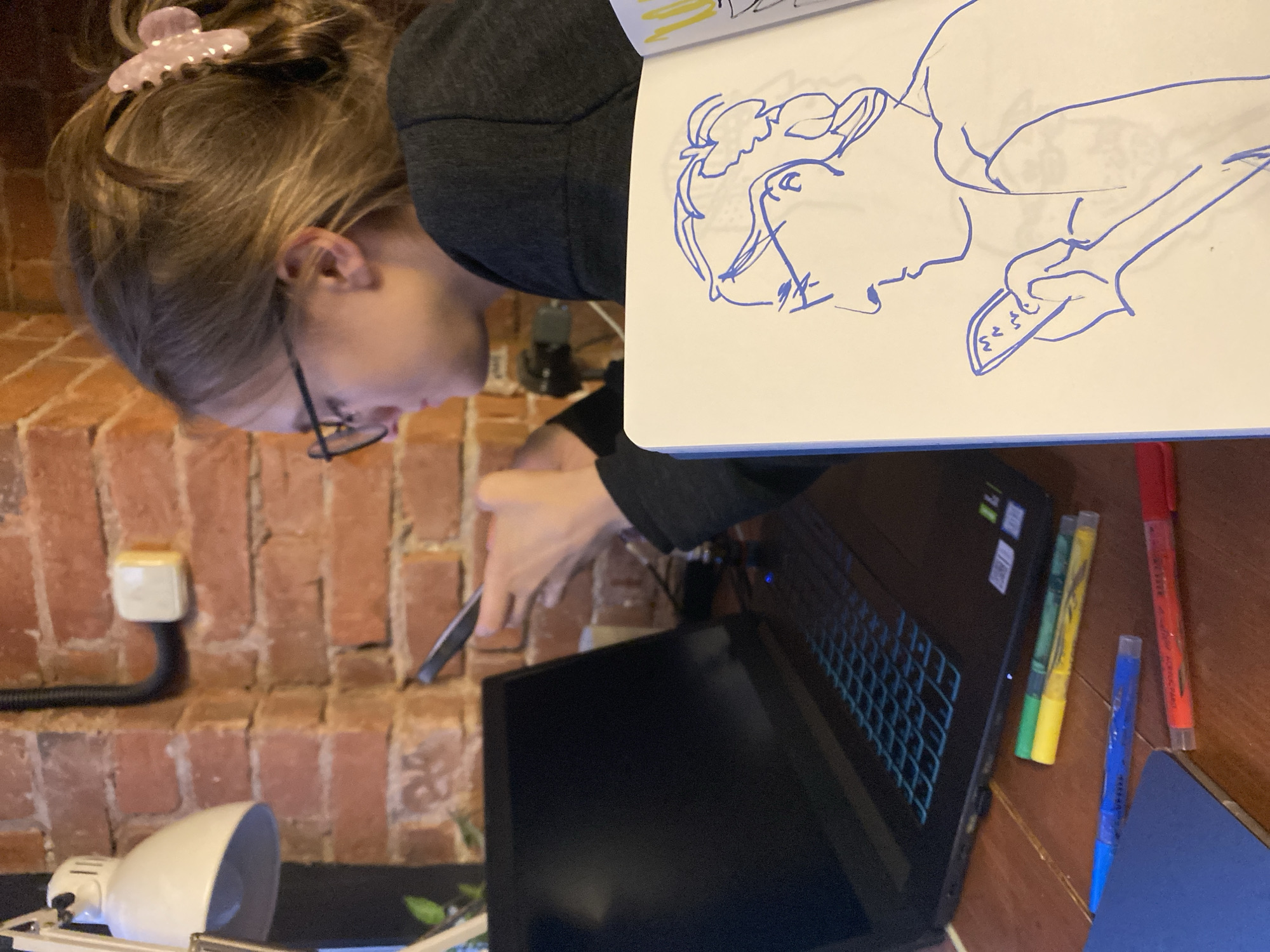 A person with blonde hair and glasses is seated at a desk. Their hair is pulled back with a pink clip. They are wearing a dark gray sweater. The person is looking at a laptop, and their hand is holding a stylus or pen near the screen. To the right of the laptop, there are several colorful markers on the desk. The person is holding a sketchbook on top of the laptop and drawing on the sketchbook, specifically drawing an animal with a hand reaching up. In the background, a brick wall can be seen, with a light switch and a desk lamp. There is no information to suggest the image was taken in Moscow, Russia.