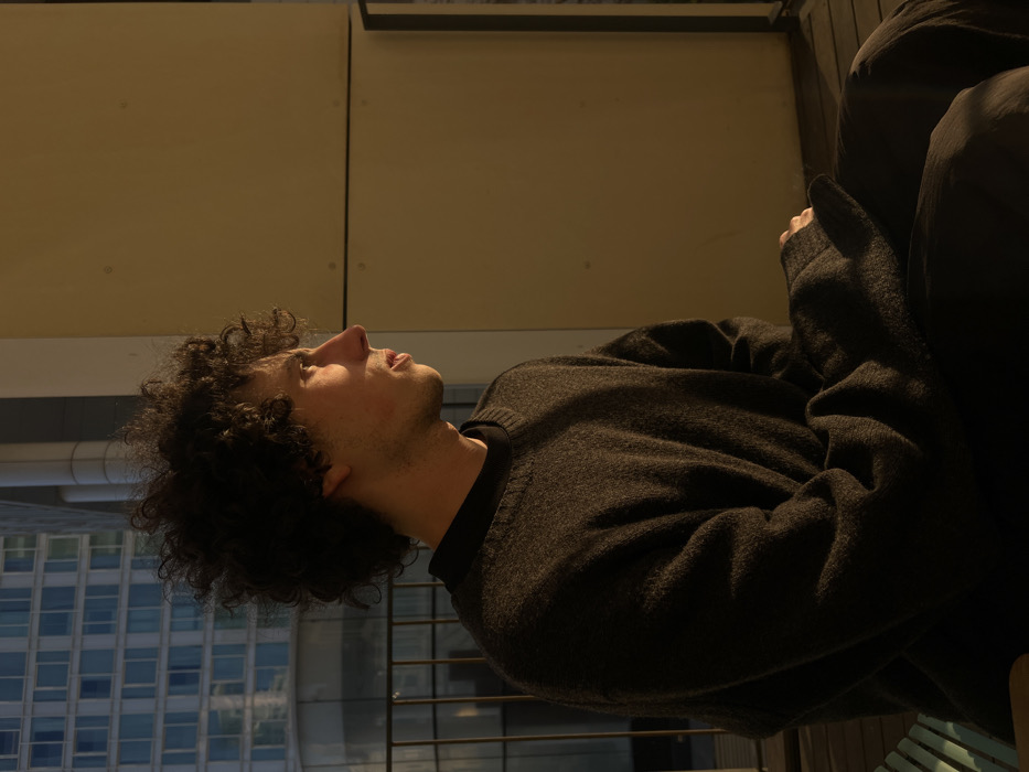 A male individual is depicted in profile, seated on an outdoor balcony or terrace within an urban environment in Tel-Aviv, Israel. He faces towards the right side of the frame, with his gaze directed beyond the visible area. The subject has dark, curly hair and is wearing a dark charcoal-grey crew-neck sweater over a dark undershirt, paired with dark trousers. Strong, warm, low-angle light illuminates the right side of his face, including the cheekbone, nose, and forehead, while the majority of his body remains in shadow. A portion of a light-colored, slatted chair or bench is visible in the lower-left foreground. Behind the individual, a dark metal railing borders the balcony. Beyond the railing, a modern high-rise building with a reflective, grid-patterned facade is visible. To the right of the subject, a flat, light beige wall panel forms part of the balcony's structure. The balcony floor consists of dark wooden slats.