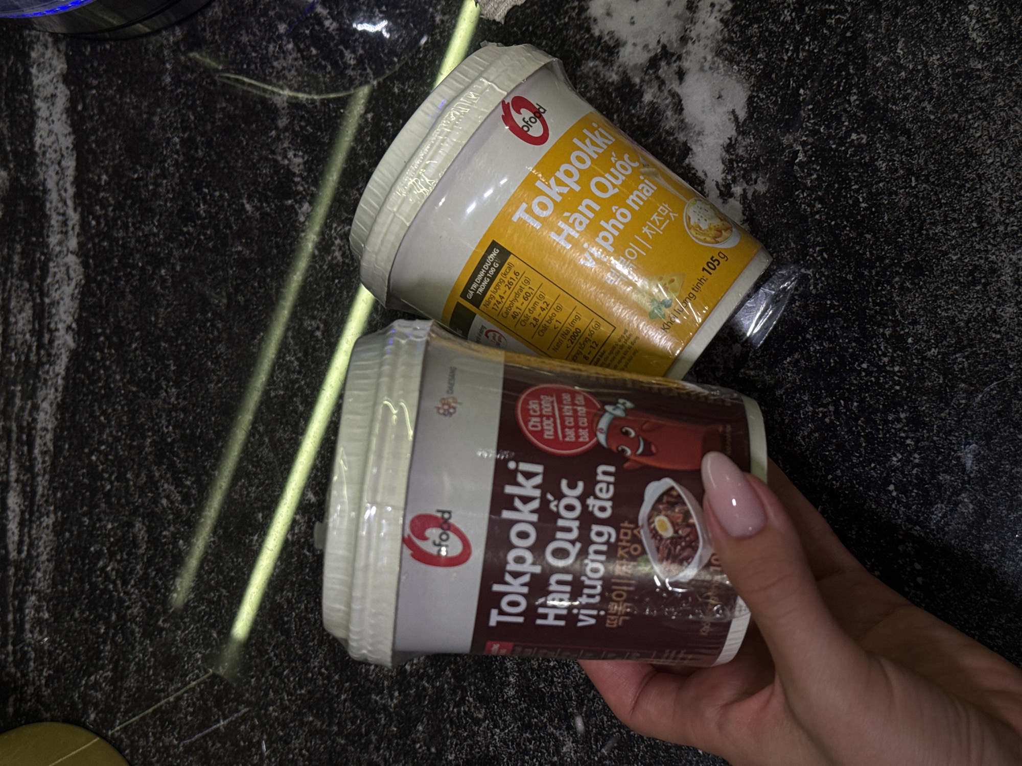 The image shows two unopened containers of "Tokpokki Han Quoc" instant food product. One container is yellow and labeled "pho mai," while the other is brown and labeled "vi tuong den". The containers are on a dark, patterned surface with a hand holding the brown container. The picture seems to be taken indoors under artificial light.