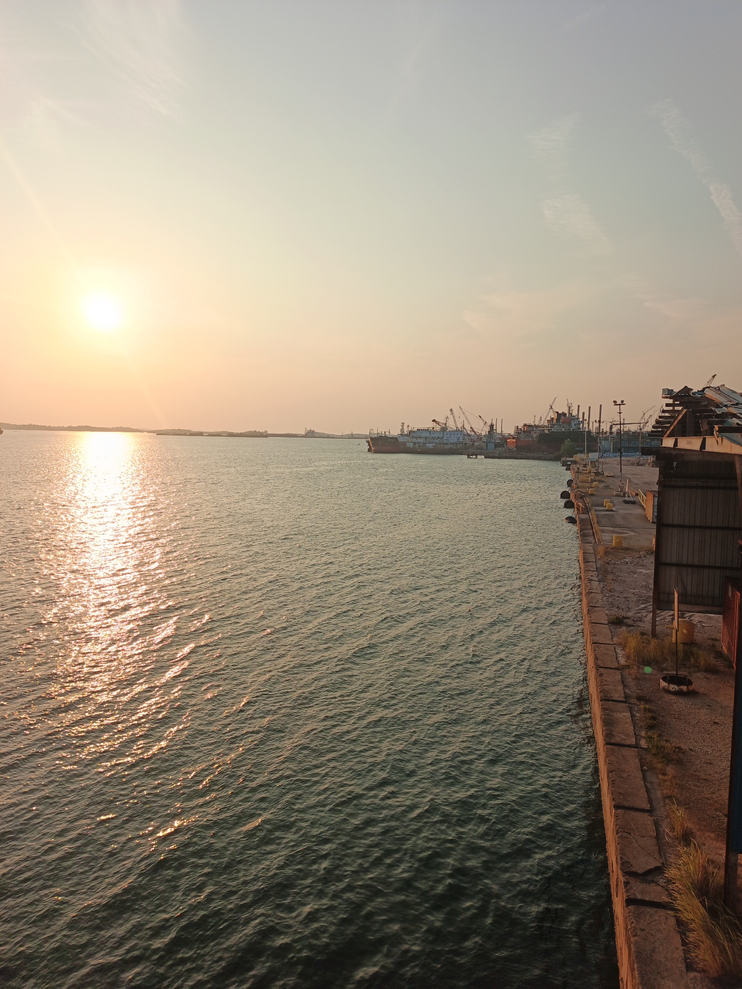The image depicts a harbor scene in Batam, Indonesia, likely during sunset. The sun is setting on the left side of the frame, casting a warm golden glow across the water and the sky. The water is calm with gentle ripples, reflecting the sunlight. Several ships, including cargo ships and tugboats, are anchored in the distance, with cranes visible on the docks. On the right side of the image, a concrete pier is visible, with some overgrown grass and yellow bollards. The sky is a soft gradient of orange and pale blue, with wispy clouds. The overall atmosphere is tranquil and serene, characteristic of a peaceful harbor at dusk.