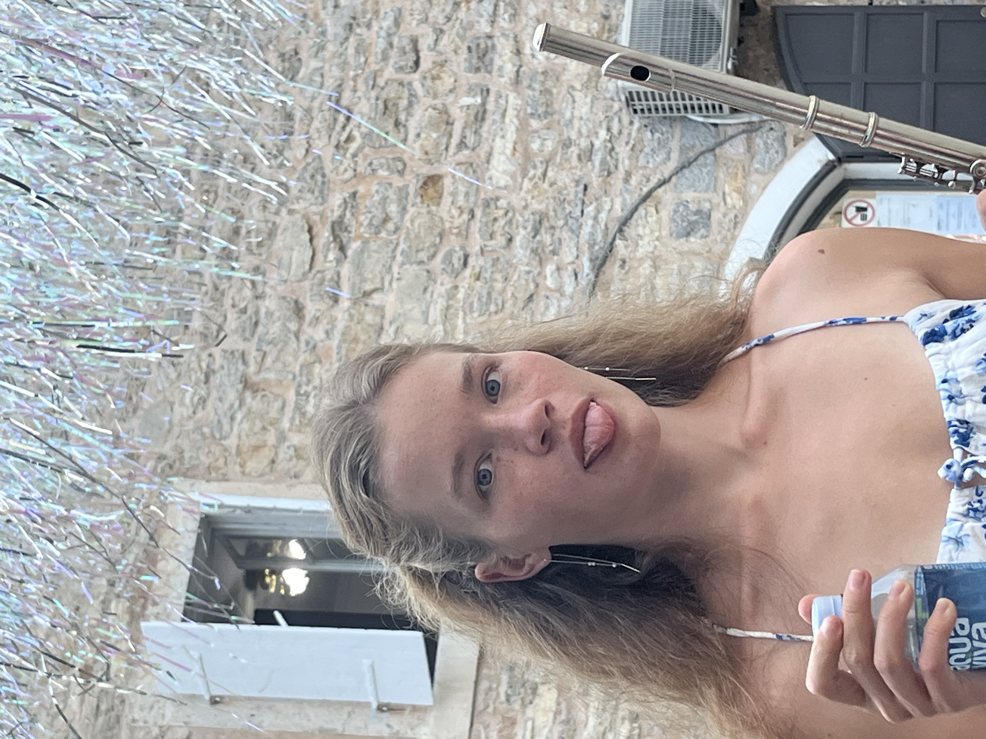 A young woman with blonde hair is pictured in Budva, Montenegro, holding a bottle of Aqua Viva water in her left hand and a flute in her right. She is wearing a white dress with a blue floral pattern and has her tongue sticking out. The background features a stone building with a window and shutters, as well as some shiny streamers hanging overhead. An air conditioning unit is visible on the wall of the building.