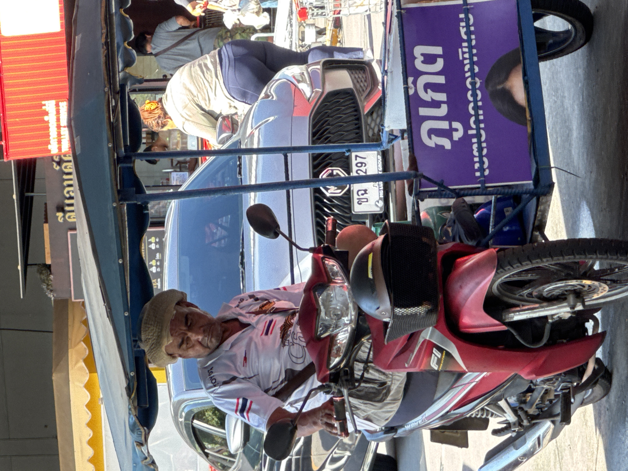 The image shows a man in a straw hat sitting on a red motorcycle attached to a cart. The man is wearing a white shirt with a red and blue stripe design. The cart has a blue canopy and a purple sign with white lettering that appears to be in Thai. The motorcycle is parked on a street in front of a gray car. In the background, there is a building with a red awning and some yellow awnings. The scene appears to be in a busy urban area. Based on the license plate of the car and the style of the motorcycle and cart, the location is likely Wichit, Thailand.