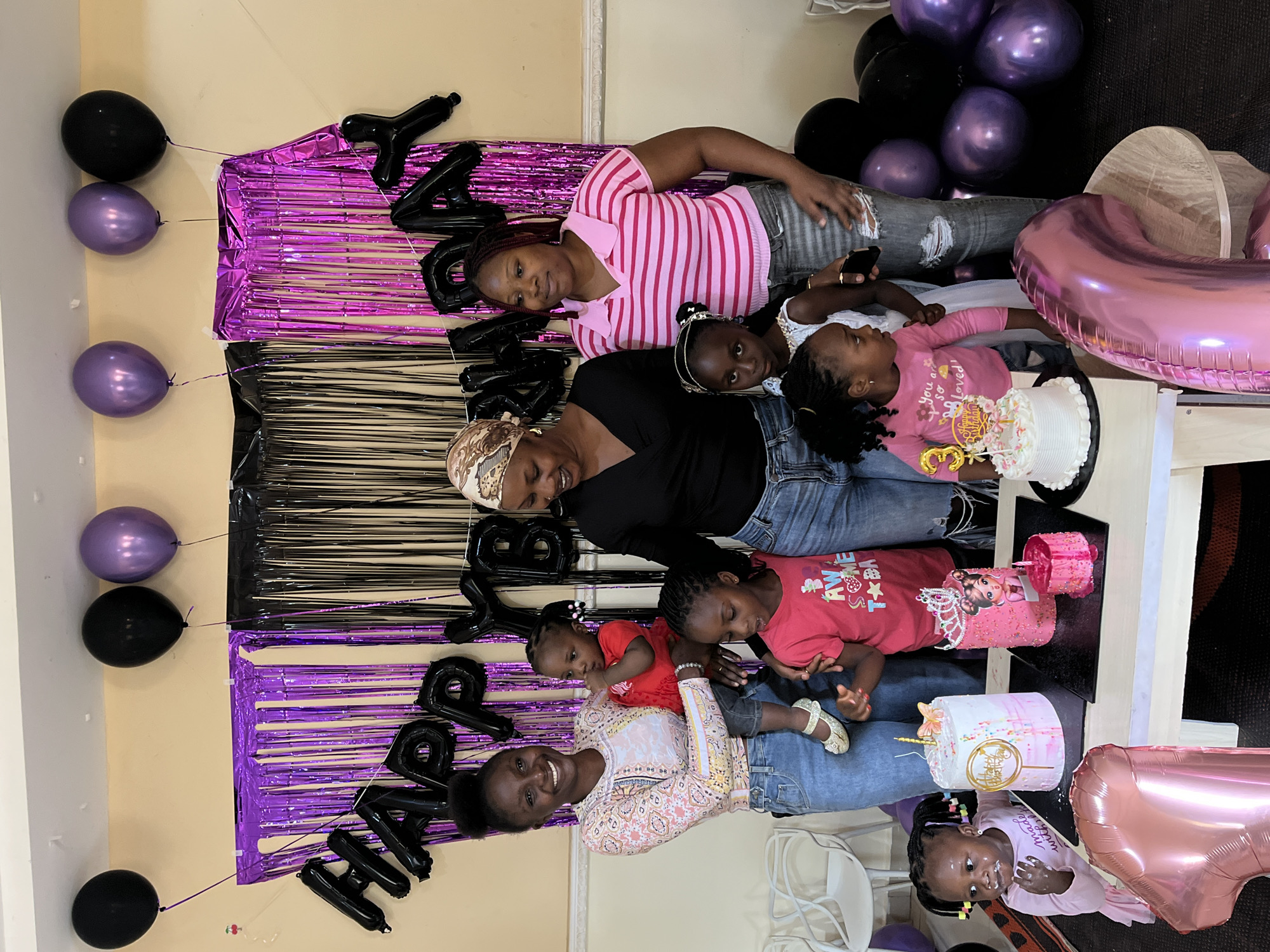 The scene depicts an indoor birthday celebration taking place in Kuru Karama, Nigeria. The background features a light yellow wall adorned with black and purple metallic fringe streamers, over which black foil balloons spell "HAPPY BIRTHDAY." Black and metallic purple balloons are visible near the ceiling and clustered on the floor to the right. Eight individuals are present, primarily adult women and young girls. 1. On the far left, a woman with dark skin and black hair in a bun wears a light-colored, long-sleeved blouse with a patterned design (pink, yellow, blue, and white) and light blue denim jeans. She holds a dark-skinned toddler wearing a red long-sleeved top with ruffles and light-colored shoes. The toddler has one hand near its mouth. 2. To the immediate right of the woman holding the toddler, a young girl with dark skin and braided hair wears a red short-sleeved t-shirt with a graphic that includes the word "AWESOME." 3. Behind a table of cakes, a woman with dark skin wears a patterned headwrap in beige and brown tones, a black long-sleeved v-neck top, and dark blue denim jeans. She exhibits a subtle facial expression. 4. To the right of the woman in the headwrap, another woman with dark skin and reddish-brown braided hair wears a pink and white striped short-sleeved collared shirt and dark grey ripped denim jeans. 5. Immediately in front of the table on the far left, a young girl with dark skin and braided hair with colorful bands wears a light pink long-sleeved shirt and has white frosting residue on her mouth and chin. She appears to be interacting with a cake. 6. Behind this girl and to the left of the cakes, a young girl with dark skin and braided hair, wearing a white sleeveless dress with a white headband, is partially visible. 7. To the right of the cakes, another young girl with dark skin and braided hair wears a pink long-sleeved t-shirt with "You are so loved!" and a bunny graphic. She stands near the edge of the table. A white rectangular table in the foreground displays four birthday cakes: * The leftmost cake is a two-tier round cake with a white top and a pink/white ombré bottom, adorned with colorful sprinkles, a gold "Happy Birthday" topper, a gold butterfly, and one candle. * Next is a single-tier round pink cake featuring a pictorial depiction of a