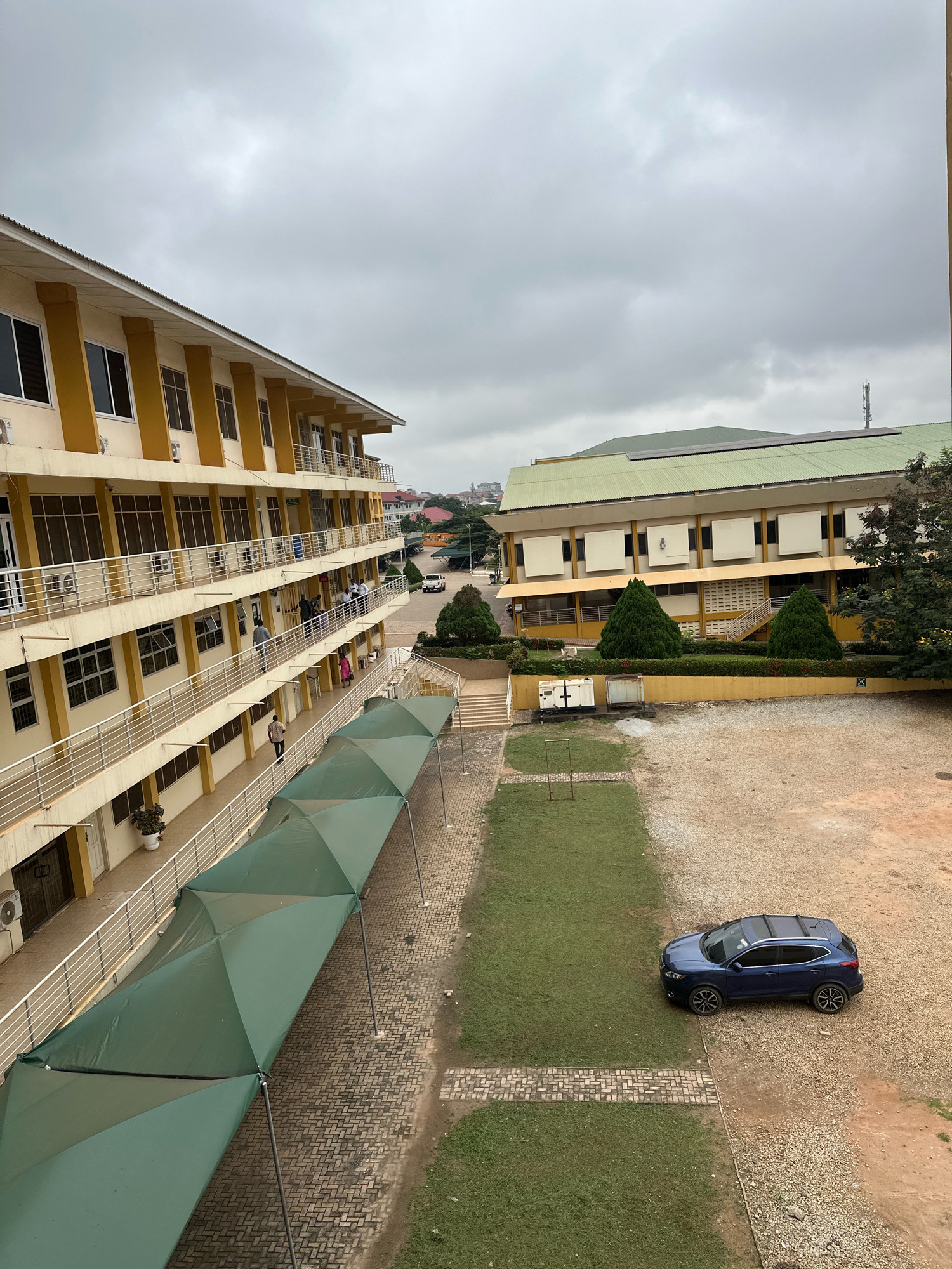 This image depicts an outdoor scene on an overcast day, likely within an institutional setting such as a school or university campus, in Kumasi, Ghana. The perspective is from an elevated vantage point, looking down and across a courtyard.
To the left, a multi-story building with a yellow facade and white railings lines a walkway covered by a series of green tensile fabric shades. The building features numerous windows and air conditioning units. Several individuals can be seen on the balconies and walkways of this building.
In the center and right of the image, another building with a green roof and yellow accents is visible. This structure has a more utilitarian appearance, with some sections featuring white louvers. In front of this building, there is a manicured lawn area bordered by gravel.
A dark blue SUV is parked on the gravel area to the right, partially on the lawn. Further in the background, other buildings and trees are visible, as well as a telecommunications tower. The overall environment suggests an urban or semi-urban academic or administrative complex.