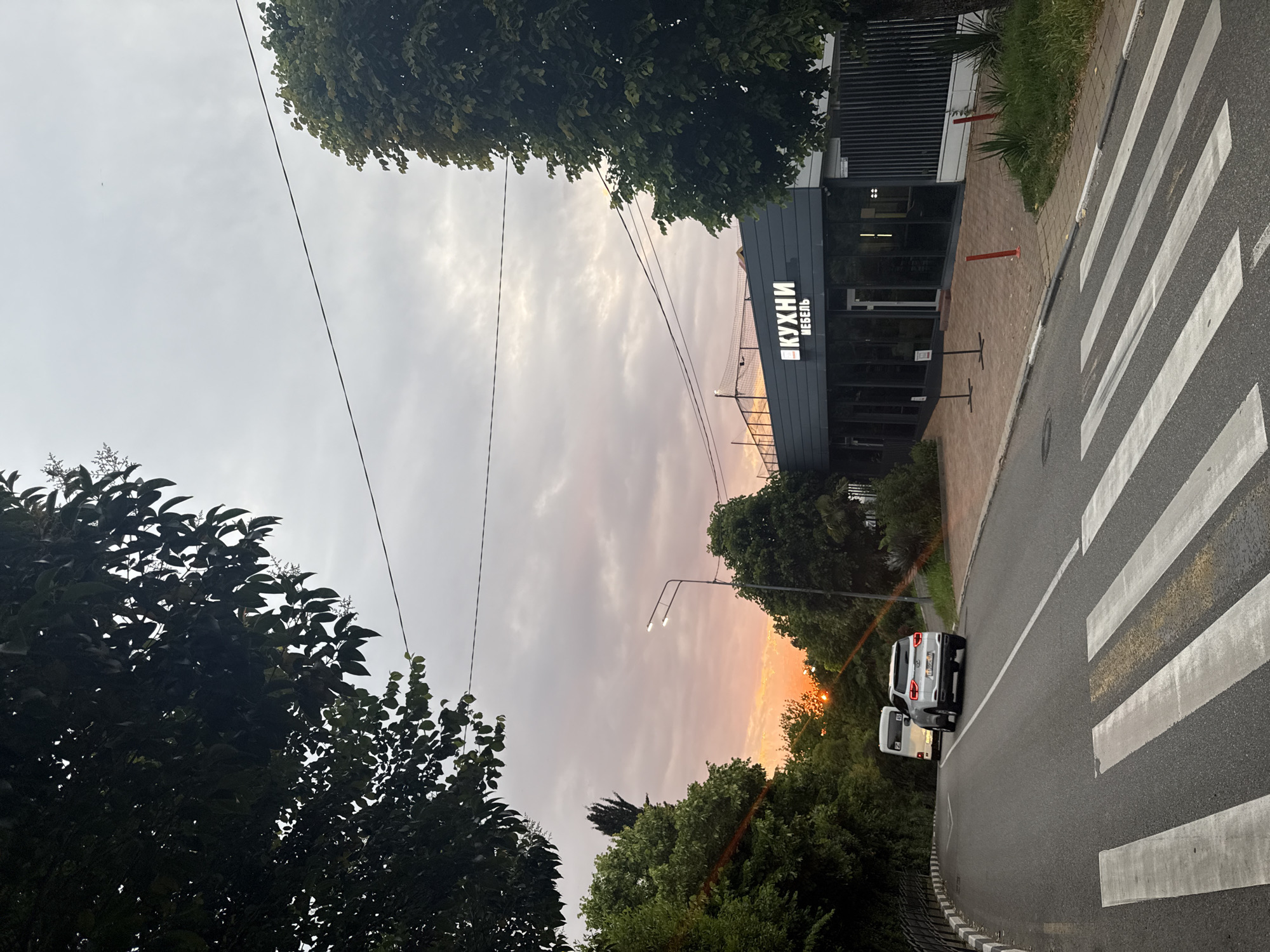 The image depicts a street scene in Sochi, Russia, during what appears to be sunset or twilight. The sky is overcast with clouds tinged with orange and pink hues from the setting sun, which is partially visible behind trees on the left side of the frame.
In the foreground, a pedestrian crossing is marked on an asphalt road. To the right of the crossing, a sidewalk paved with reddish-brown tiles runs alongside a building. The building is modern, with a dark-grey facade and large glass windows. Above the entrance, illuminated white text in Russian reads "КУХНИ" (Kitchens) and "МЕБЕЛЬ" (Furniture). Red poles are strategically placed along the sidewalk in front of the building.
On the road, two vehicles are visible. A white minivan is driving away from the viewer and upwards on the curving road. Following behind it is a white SUV, also heading away. The road is bordered on the left by a low concrete barrier with a decorative edge, and then by a slope covered with lush green trees and foliage. More trees and vegetation are also present on the right side of the street, partially obscuring the view of the building and the sky. Power lines are strung across the upper portion of the image. A street lamp with two lights is positioned on the right side of the road, illuminating the area. The overall impression is a tranquil urban environment transitioning into evening.