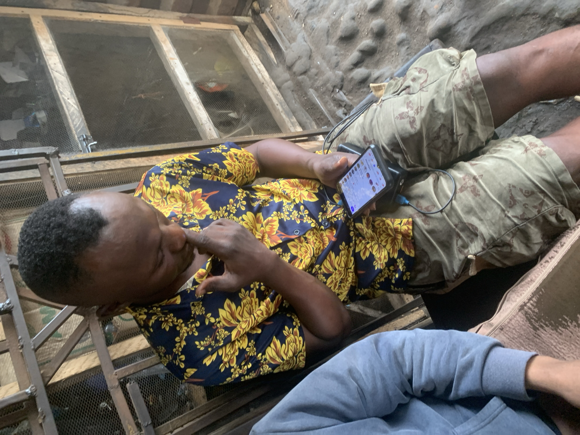 A close-up, eye-level shot shows a Black man wearing a blue and yellow floral shirt and camouflage shorts. He is seated, with his left hand touching his chin as he looks down at a smartphone resting on his lap. The phone is plugged into a black power bank with a blue charging cable. He appears to be in an outdoor or semi-outdoor environment, possibly a workshop or an area with caged structures behind him. The ground is rough and uneven. Another person's arm, wearing a blue hooded sweatshirt, is visible on the left side of the frame. The scene suggests a casual moment of using technology amidst a rustic setting in Shagamu, Nigeria.