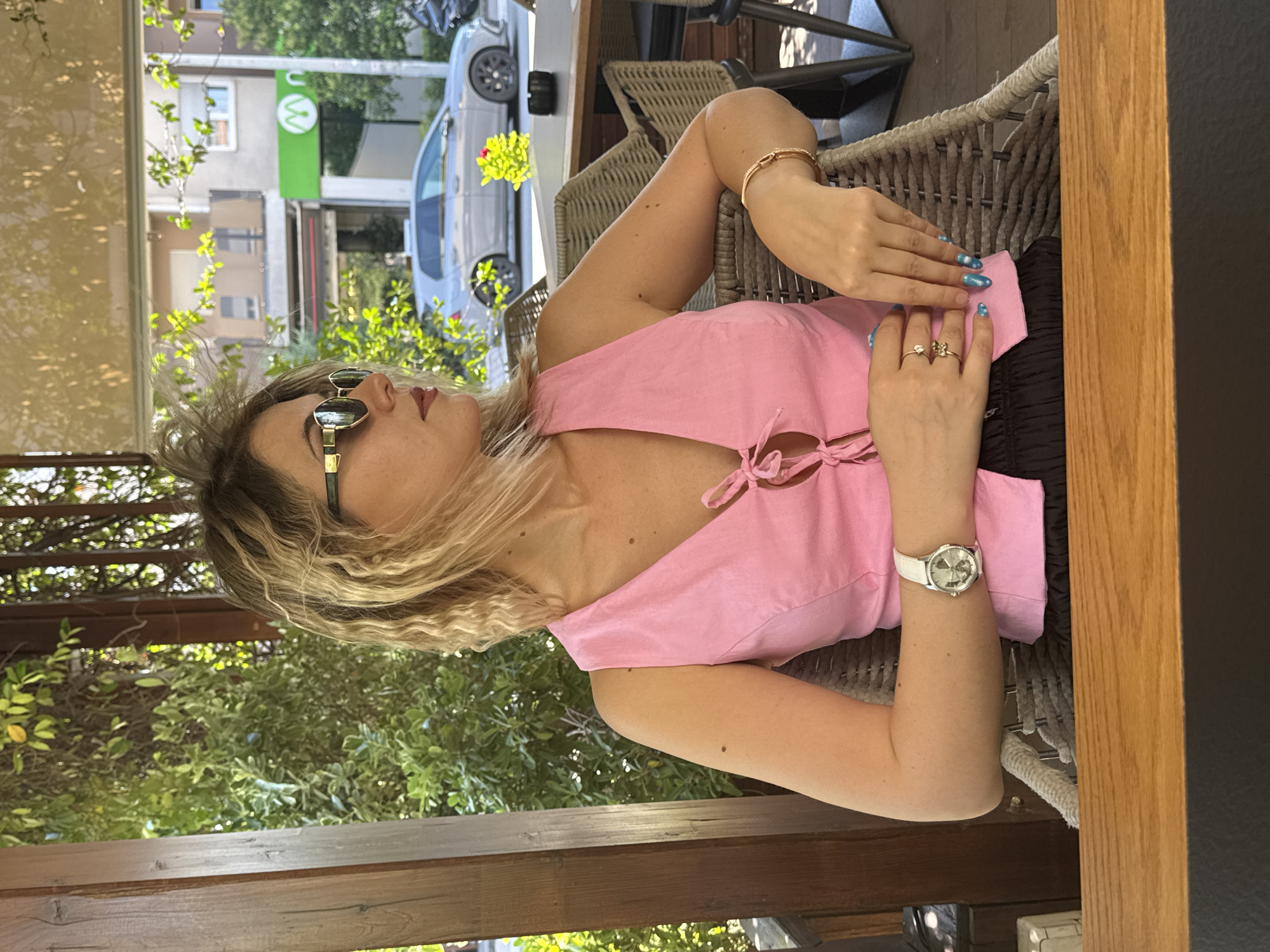 A woman with blonde, wavy hair is seated at an outdoor cafe table, looking to her right. She is wearing large, rectangular sunglasses with gold frames, a pink sleeveless V-neck top with tie closure at the bust, and a black skirt. She has a white-faced watch on her left wrist and several rings on her fingers, including one with a small diamond. Her fingernails are painted with a blue and white swirling design. Her right arm rests on the table, and her left arm is bent with her hand resting on her lap. Behind her, there are wicker chairs, a wooden table, and lush green foliage. In the background, a silver car is parked on a street in front of a building with a green sign that has a white "M" logo. The scene is illuminated by natural sunlight.
