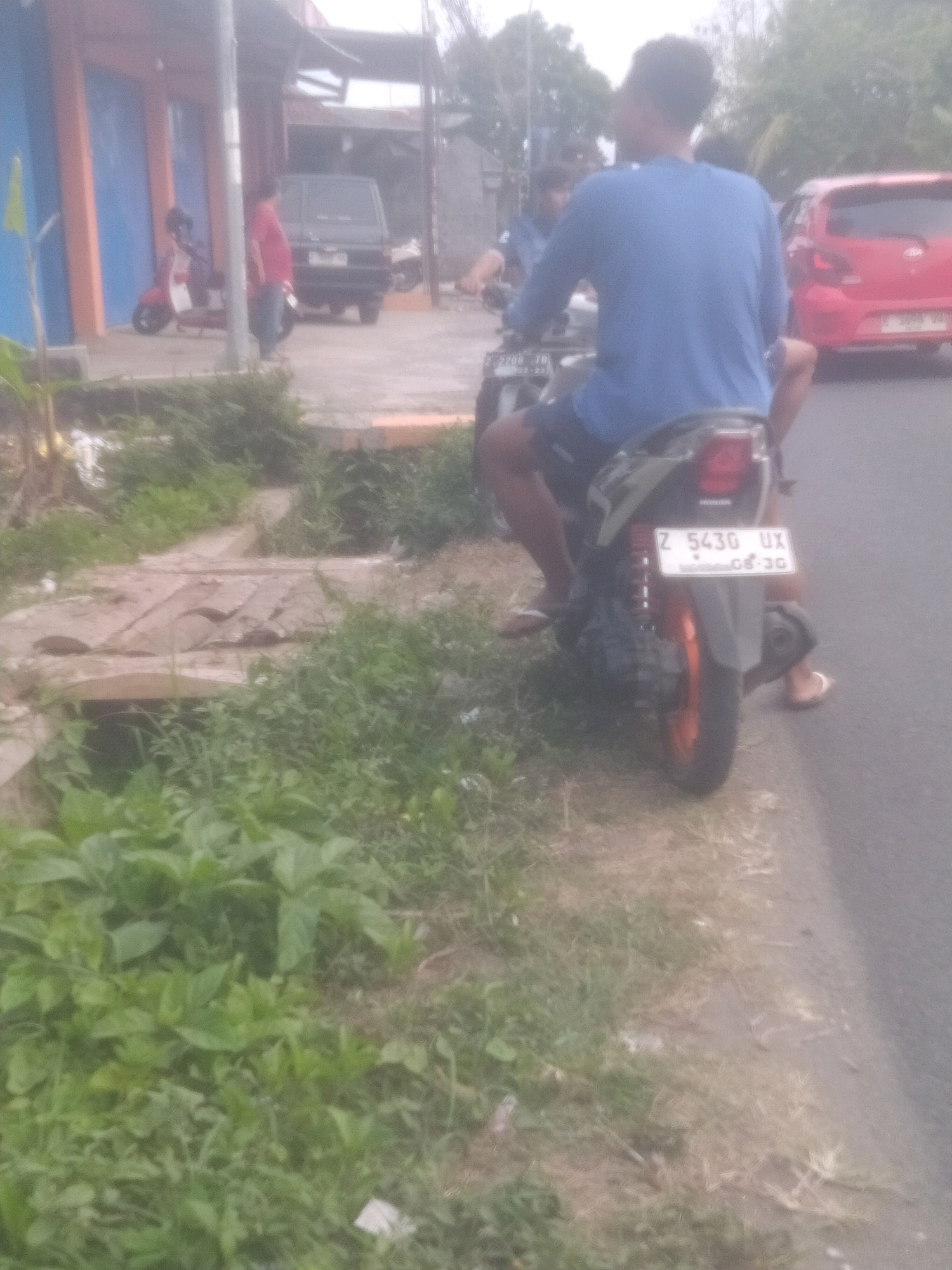 In the city of Parigi, Indonesia, this image captures a street scene. Two individuals are present, both on a motorcycle. The person in the foreground, wearing a blue long-sleeved shirt and dark shorts, is seated on the motorcycle. Their back is to the camera. Another person is partially visible in front of them, also on the motorcycle. The motorcycle has a license plate that reads "Z 5430 UX" with "08-26" below it. The motorcycle is on a paved road, with a grassy area and a small wooden bridge to the left. In the background, a dark-colored van is parked, and further back, a red car is visible. A building with an orange and blue facade stands on the left side of the street, and a red scooter is parked in front of it. The scene is outdoors, with trees and foliage in the background, suggesting a moderately developed urban or suburban environment.