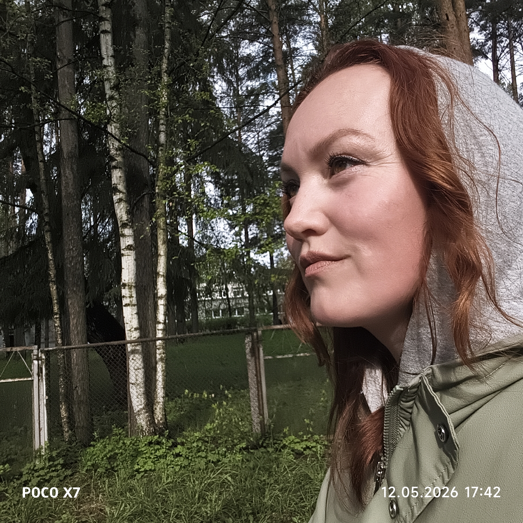 A photograph shows a fair-skinned woman with red hair, wearing a gray hooded sweatshirt and an olive-green jacket, standing outside in a wooded area. The trees behind her include birch, with white bark visible, and other unidentified trees. A chain-link fence and grass are also visible. The date "12.05.2026 17:42" is printed on the lower right corner, and "POCO X7" on the lower left. The photograph was taken in Chaikovsky, Russia.