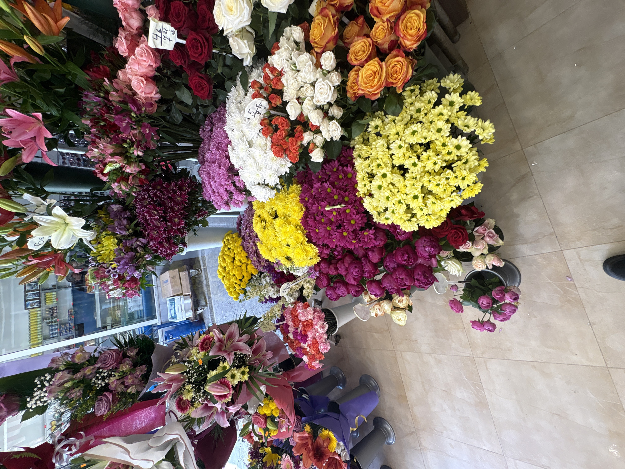 The image shows a flower shop with a variety of flowers on display. The flowers are in different colors, including red, pink, white, yellow, and orange. Some of the flowers are in vases, and others are on shelves. There are people and other objects that can be partially seen through the glass of the store. The location appears to be in a city, possibly Sofia, Bulgaria.