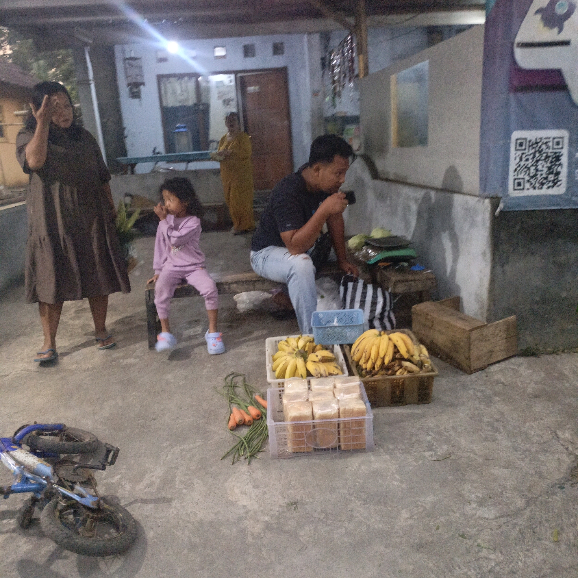 In Salatiga, Indonesia, a street scene unfolds with several individuals and objects indicative of a market or roadside stall. A woman in a dark brown dress stands to the left, gesturing with her right hand. Beside her, a young child in light purple attire sits on a low wooden structure. Further back, a woman in a yellow outfit is visible, appearing to be involved in the scene. In the foreground, a man in a black t-shirt and light blue jeans is seated, holding a dark object to his face, possibly eating or drinking.
The ground is paved with concrete, and several items are displayed for sale. Two baskets of bananas are prominent, one white and the other brown. Next to the bananas, a white plastic crate contains an assortment of packaged white bread. To the left of the bread crate, a bundle of long green beans and several orange carrots are laid out on the ground. A blue bicycle lies on its side in the lower left portion of the image. In the background, a building with windows and a doorway is visible, with a colorful advertisement featuring a rocket and a QR code on the right side. The lighting suggests it is either dusk or early morning.