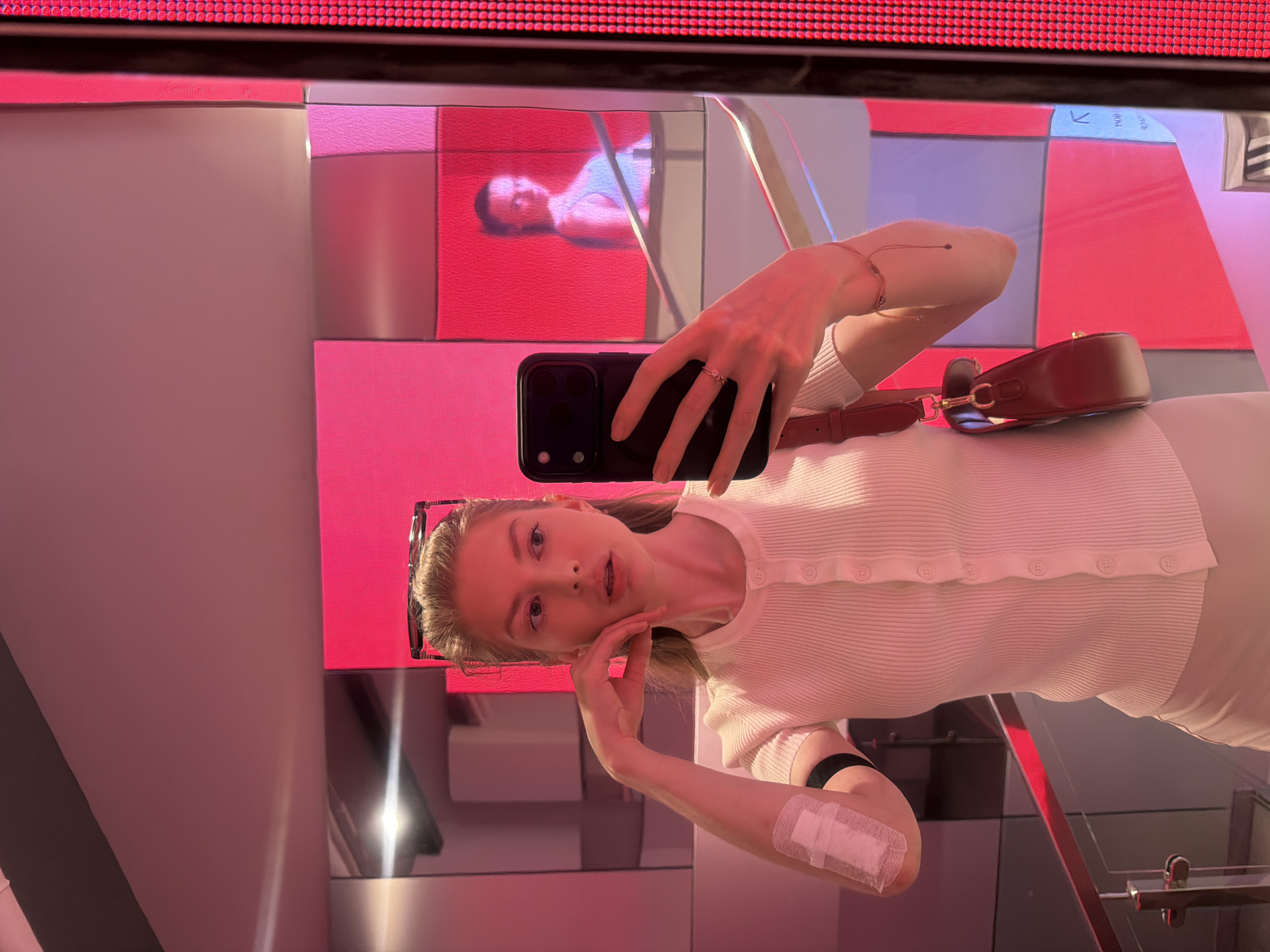 A woman is taking a selfie in a mirror. The mirror is in a modern, brightly lit interior with pink, white, and silver panels on the walls. The scene is illuminated by the reflections on the mirror. The woman is fair-skinned with blond hair and is wearing a white top with buttons. She is holding a black smartphone in her right hand, and her left hand is near her face. She has sunglasses perched on her head and a brown purse with a strap over her shoulder. There is a white bandage on her left arm and a black bracelet on her left wrist. A reflection of another person is visible in the mirror. The reflection shows Moscow, Russia.