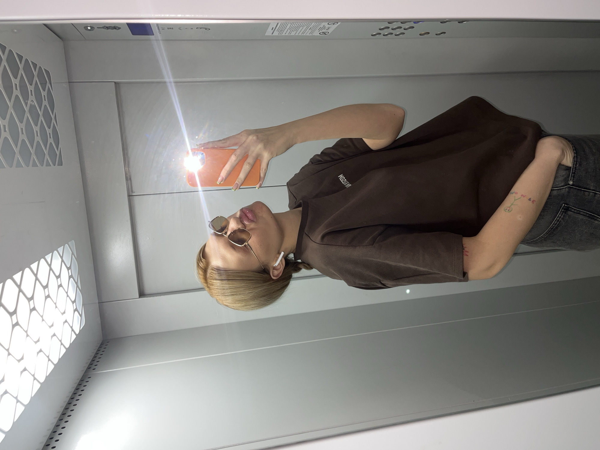 A person with blonde hair and sunglasses takes a selfie in an elevator with an orange smartphone. The person wears a brown shirt with writing on it, jeans, and an earbud. The elevator has a white interior with ventilation grates and lighting. The location is the city of Chaikovsky, Russia.