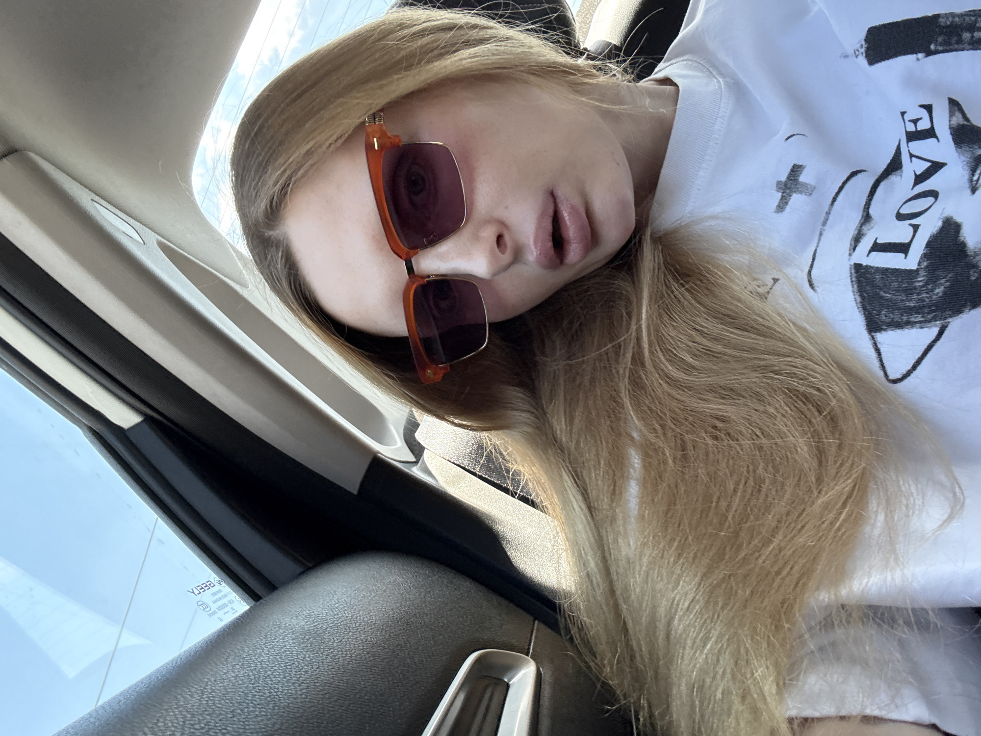 A young woman with long blonde hair, wearing sunglasses and a white t-shirt with a graphic print, is seated in the back of a vehicle. The interior of the car, including the black leather seat, the headliner, and a portion of the side window, are visible. The woman is looking towards the camera, and her mouth is slightly open. The environment appears to be daytime, with natural light entering the car.