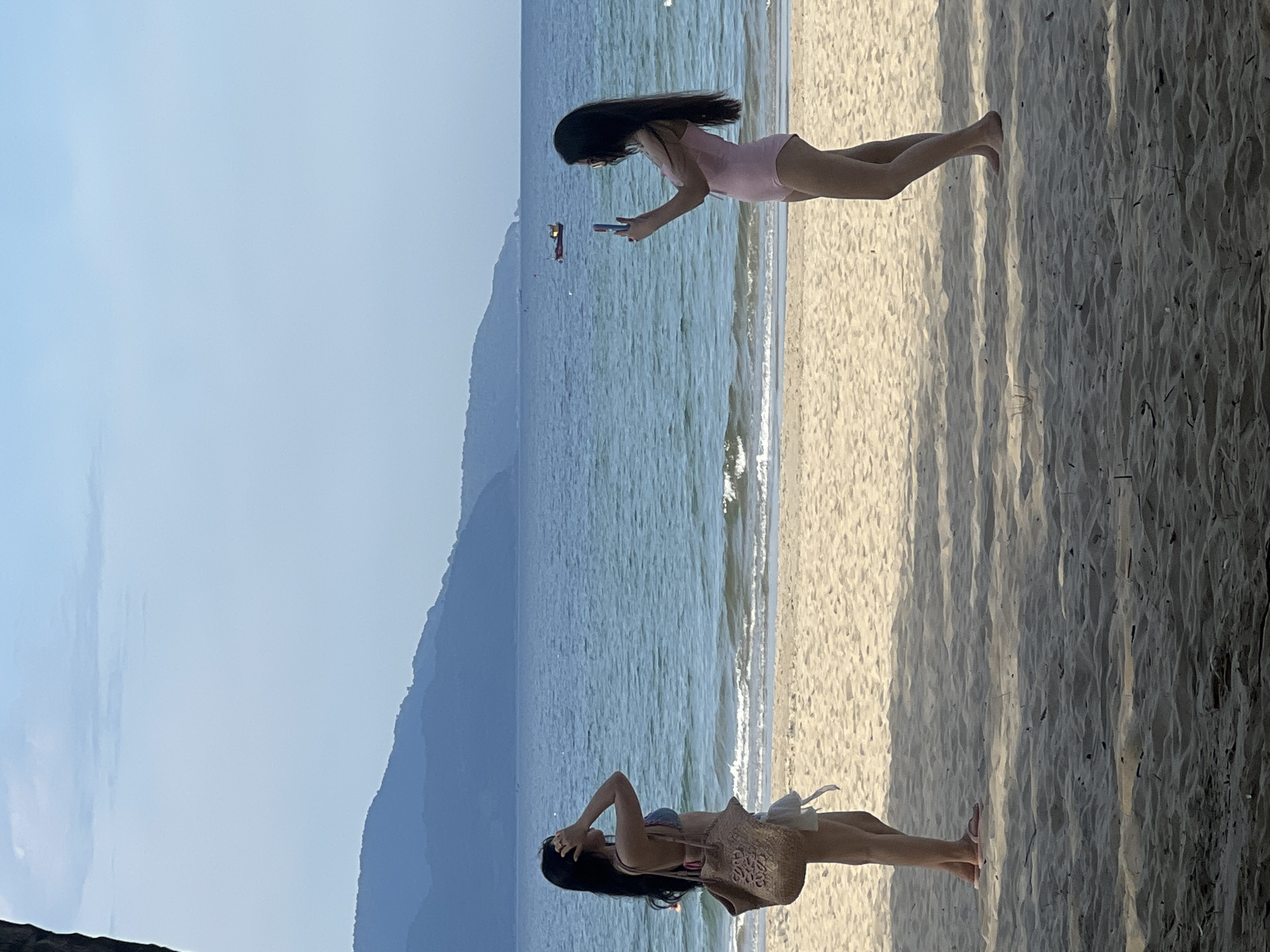 Two women are on a sandy beach in Da Nang, Vietnam, with the ocean and mountains in the background. The woman on the left, wearing a bikini and shorts, is posing with her hands near her head and a woven bag at her side. The woman on the right, in a pink one-piece swimsuit, is taking a photo of the woman on the left with her smartphone. The sea is blue with gentle waves, and the sky is clear with distant mountains. A small boat is visible on the water. The foreground is a sandy beach with shadows from nearby trees.