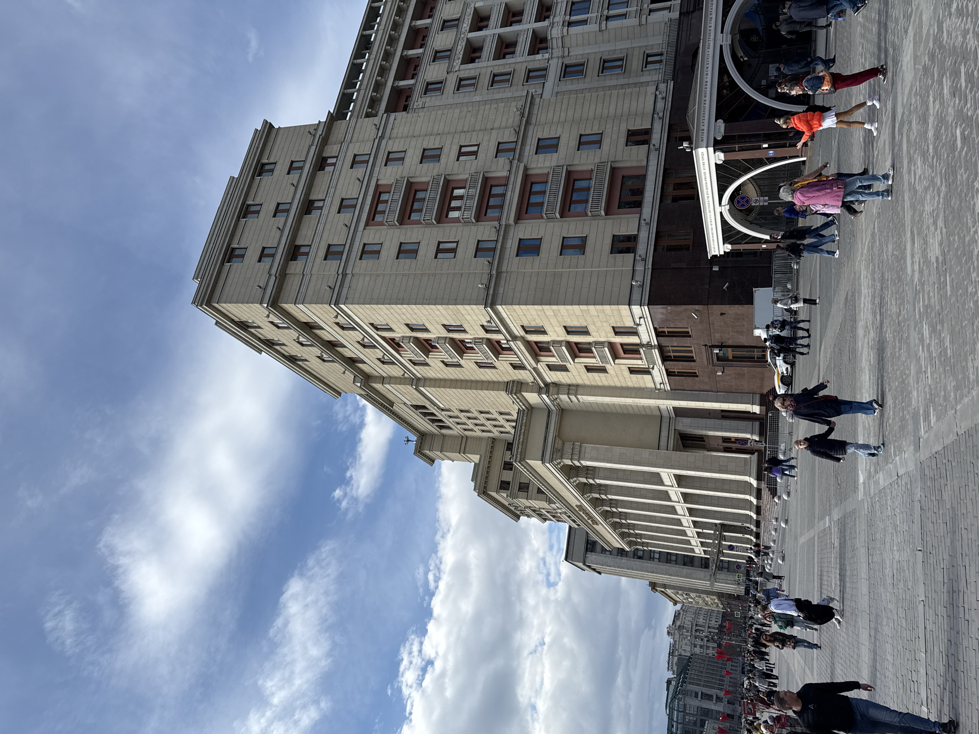The image shows a tall, beige-colored building with numerous windows, likely in Moscow, Russia. The architecture features a repetitive pattern of window placement and decorative elements. The building has a distinct crown at its top. Below the building, on a paved area, people are walking and standing. One prominent structure is a decorative archway with text in what appears to be Russian Cyrillic above it. A street runs beside the building. The sky is blue with scattered clouds. The photo is taken from a low angle, emphasizing the building's height. Flags are also visible in the distance.
