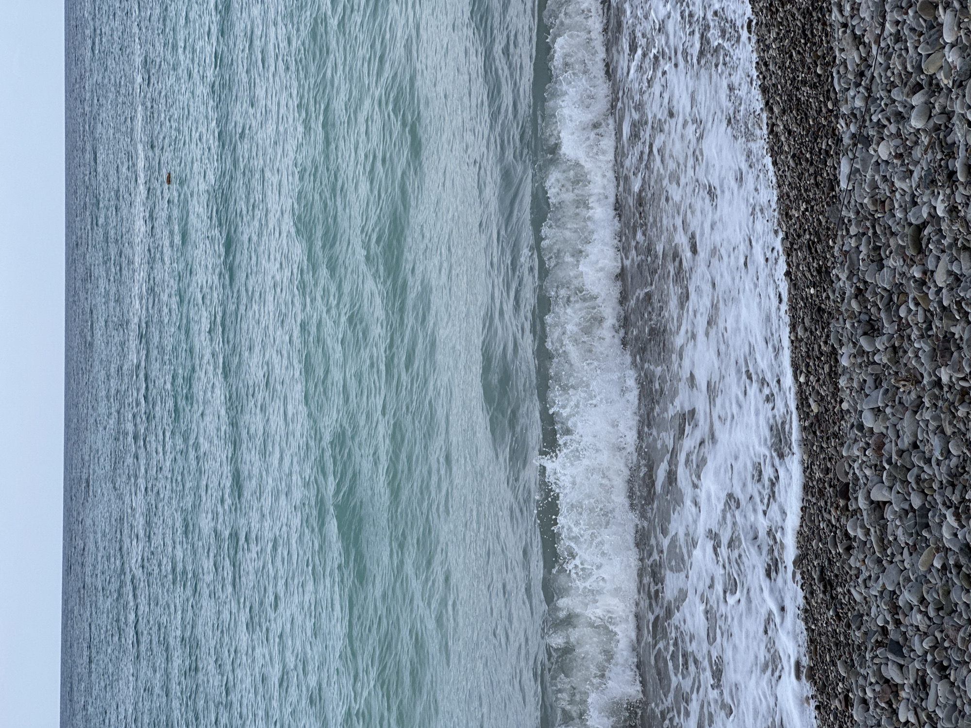 The image captures a static scene of a pebble beach meeting the sea at Šušanj, Montenegro, under an overcast sky. The upper quarter of the frame shows a pale, uniform light grey sky. The majority of the image is occupied by the sea, which exhibits a spectrum of blue-green hues, transitioning to a deeper grey-blue towards the horizon. The water surface displays ripples, and a notable wave is in the process of breaking onto the shore in the mid-ground, generating white foam and spray. A small, dark, oblong object, possibly debris or a marker buoy, is visible floating in the mid-distance on the water, slightly to the right of the frame's vertical center. The foreground consists of a pebble beach, composed of numerous smooth, predominantly grey and dark-toned stones of varying sizes, extending horizontally across the lower portion of the image and meeting the foamy edge of the breaking wave. No individuals, animals, or significant artificial structures are present within the depicted area. The lighting is diffused and consistent.