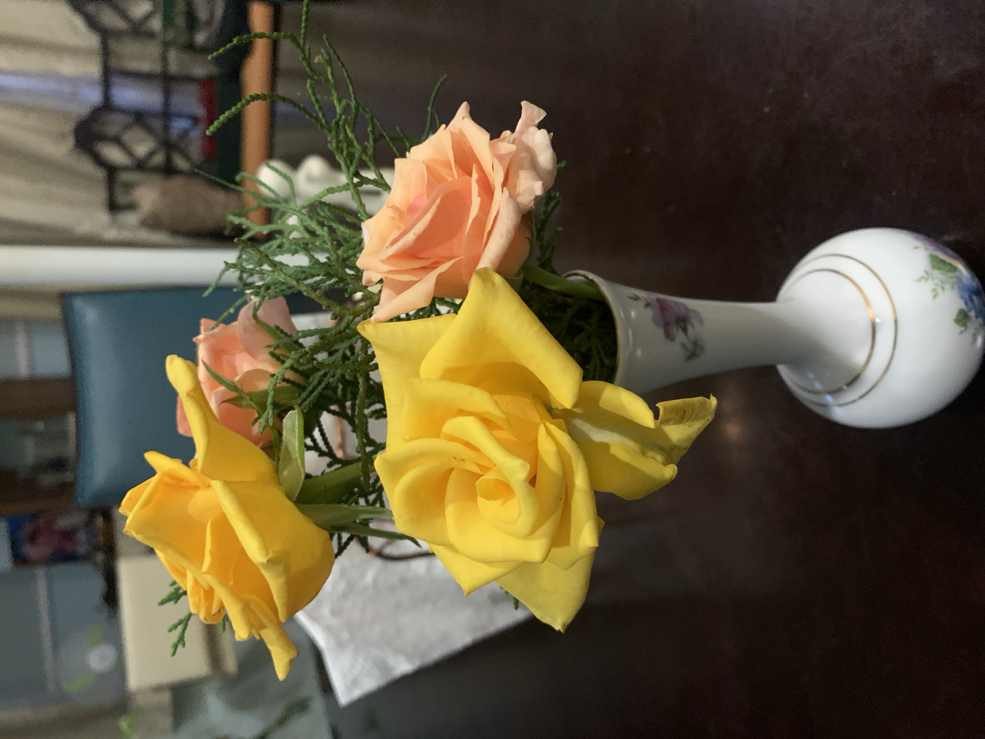 A close-up, eye-level shot captures a small vase of flowers on a dark wooden table. The vase is white with delicate gold trim and floral details. Inside, two bright yellow roses and one soft peach rose are arranged with sprigs of greenery. The background is blurred, showing hints of furniture and a window, suggesting an indoor setting. The lighting is soft, highlighting the textures and colors of the flowers. The scene is static, with no people or discernible actions. This image is from Aketu-Oja, Nigeria.