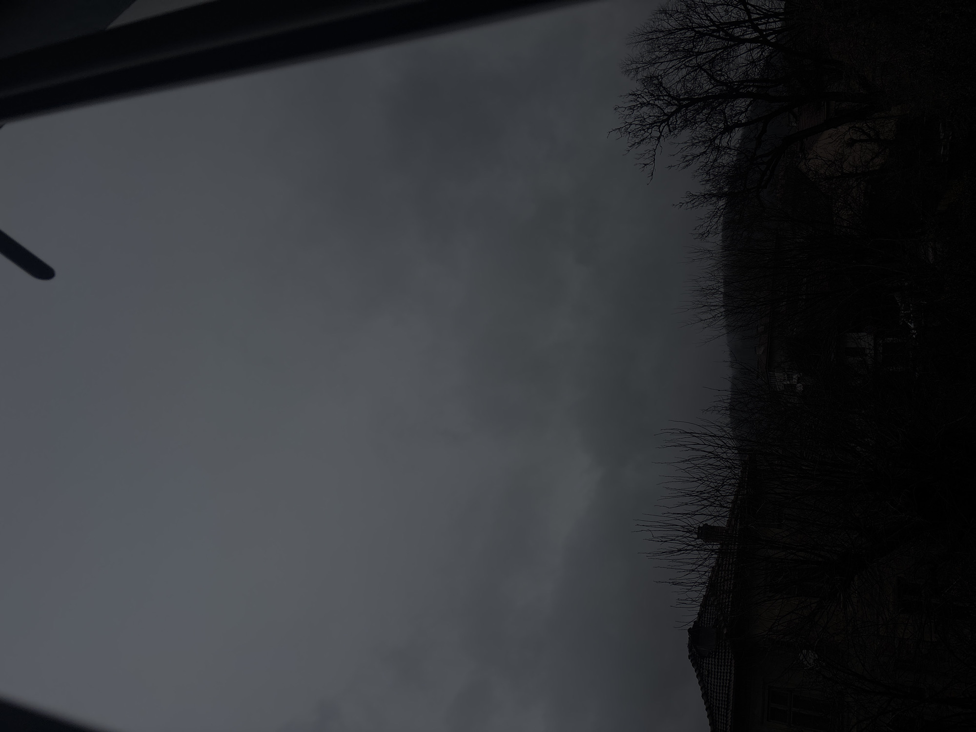 The image shows a dark, overcast sky above Troyan, Bulgaria. A window frame is visible at the top left, suggesting the image was taken from indoors. A building and bare trees are visible on the right side of the frame. The scene is dimly lit.
