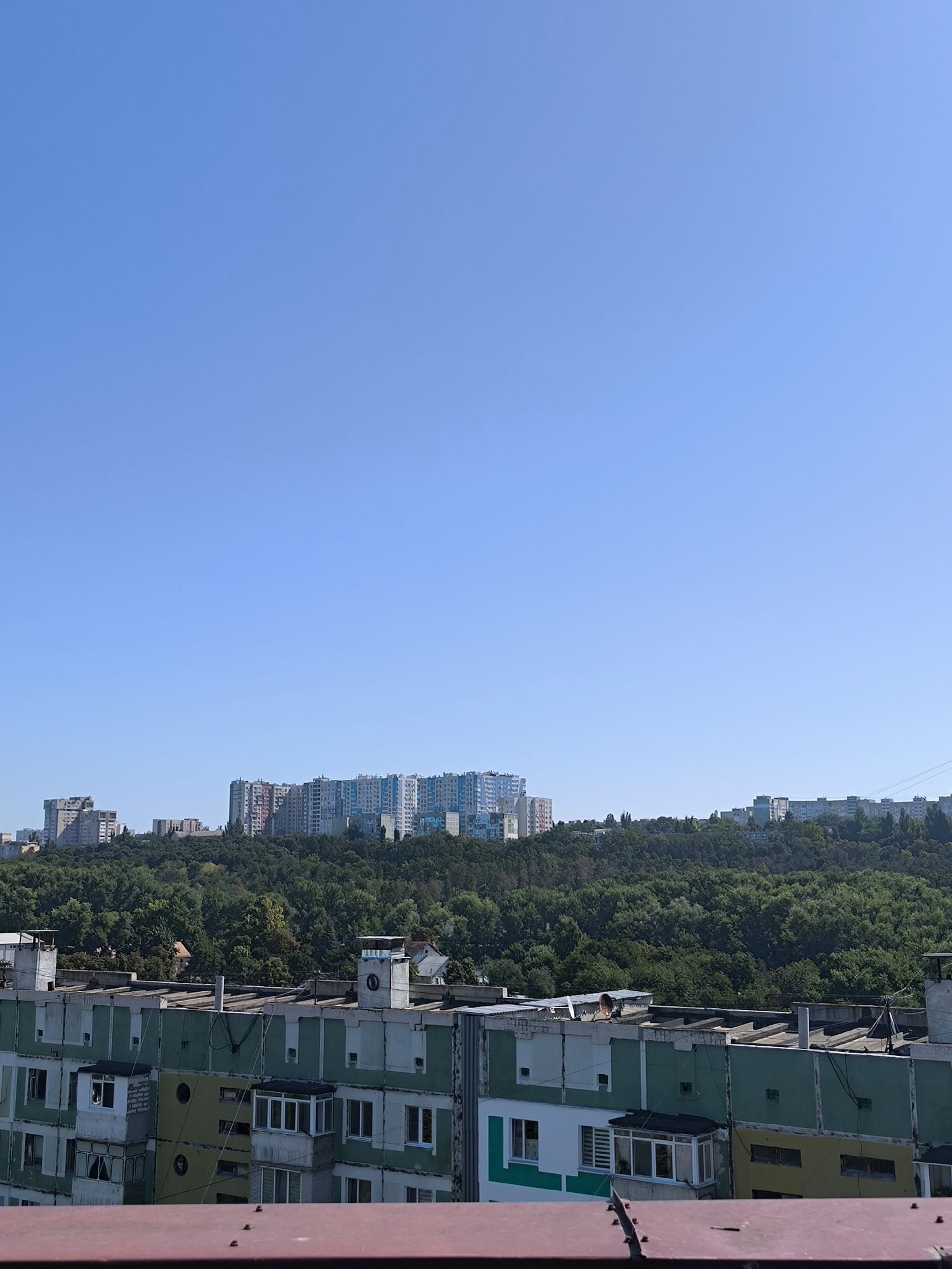 This is a daytime, eye-level, outdoor shot of a residential area, likely in Chișinău, Moldova, given the architectural style of the buildings. The foreground is dominated by the top of a weathered, reddish-brown structure, possibly a balcony railing, with visible rivets. Behind this, a multi-story apartment building stretches across the frame. Its facade is a patchwork of faded green, yellow, and white paint, with various window styles and balconies, some enclosed with glass. The building appears to be in a state of disrepair, with some windows boarded up and visible signs of age. On the roof of this building, several ventilation pipes and other structures are visible.
Beyond this foreground building, a dense, green forest covers a hillside. In the distance, atop the forested hill, a line of modern, colorful apartment buildings stands against a clear, bright blue sky. These buildings are much taller and more contemporary than the one in the foreground, featuring diverse colors like light blue, pink, and white. The contrast between the older, somewhat dilapidated apartment building in the foreground and the newer, vibrant structures in the background suggests a mix of development stages within the city. The overall impression is of a sunny day in a suburban or urban fringe area characterized by both older Soviet-era residential blocks and newer construction.
