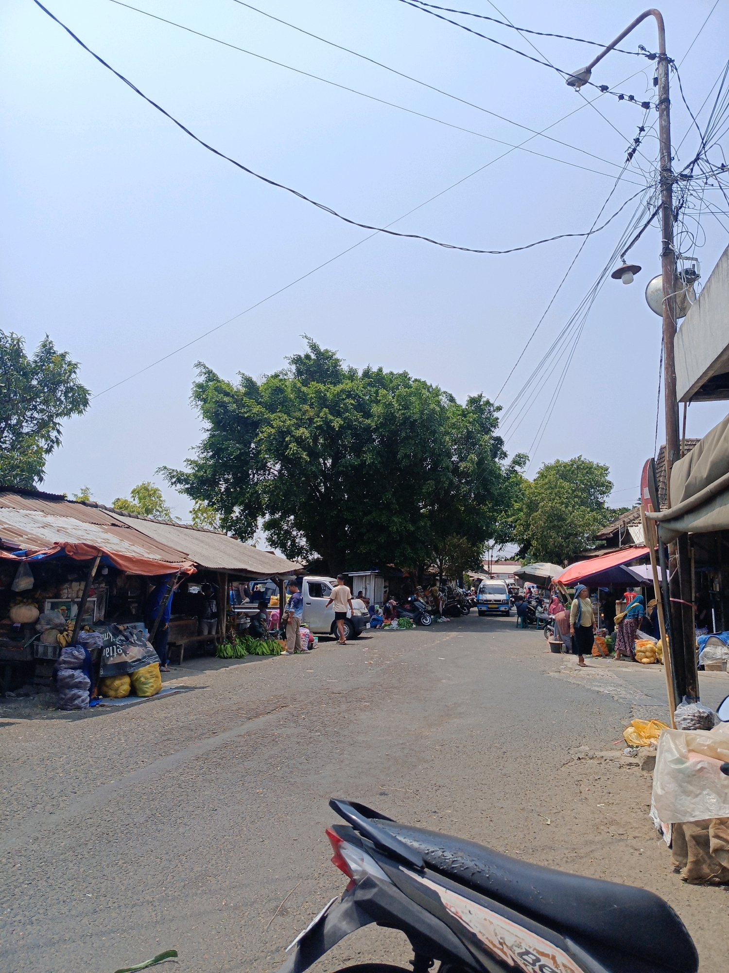 This image depicts a busy street scene at a marketplace in Sukowinangun, Indonesia. The road is paved and appears to be a thoroughfare lined with temporary market stalls on both sides. On the left, a row of stalls with corrugated metal roofs and tarp awnings display various goods, including what appear to be large bags of produce like potatoes and bananas, as well as other items. Several people are visible tending to the stalls or browsing.
Further down the street, a light-colored van is parked. On the right side of the road, more market stalls are set up, with people interacting and shopping. A woman wearing a light yellow headscarf and dark skirt is standing in front of a stall displaying sacks of yellow produce. Another stall has a red and blue umbrella providing shade.
The background features lush green trees, suggesting a somewhat rural or suburban setting within the city. The sky is bright and hazy, indicating a sunny day. Overhead, a complex network of electrical wires is strung across the scene, attached to a tall utility pole with two streetlights. In the foreground, the back of a black motorcycle is visible, with its seat and tail light in focus, suggesting the photo was taken from the perspective of someone on or near a motorcycle. The overall impression is one of a vibrant, active local market.