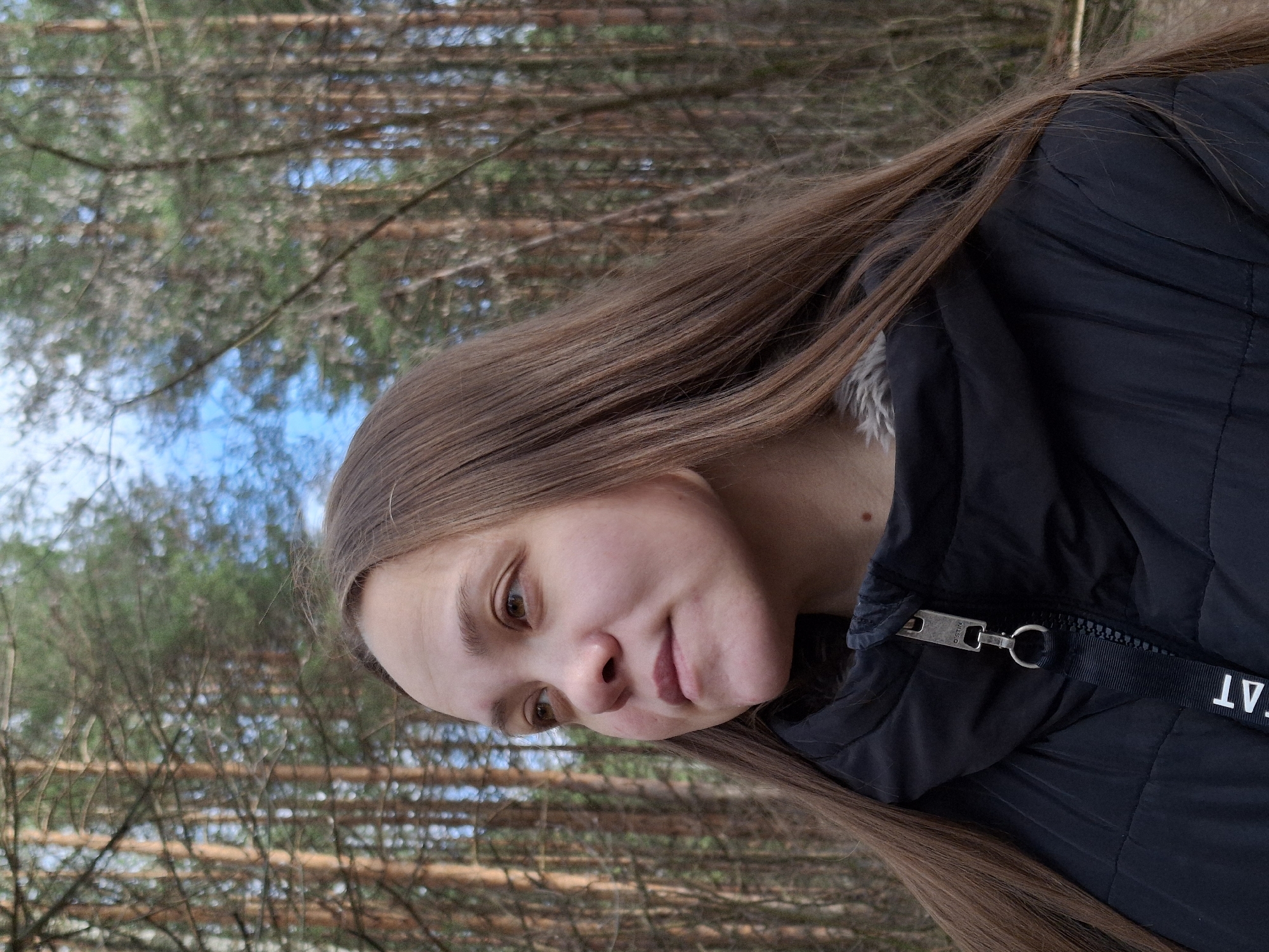 The image displays a head-and-shoulders portrait of a young woman taken outdoors, rotated 90 degrees counter-clockwise. The woman, with long, straight, light brown hair, is positioned against a blurred background of a forest.
Her face, illuminated by natural light, shows light brown eyes and a subtle, closed-mouth smile, conveying a calm expression. She is wearing a dark, possibly black, puffer jacket. A prominent silver-colored metal zipper pull is visible on the jacket, with the letters "N.B.D." faintly inscribed on it. Attached to the zipper pull is a black ribbon or fabric strip featuring white text, partially legible as "AT". A light-colored, fluffy collar or lining is visible inside the jacket near her neck.
The background consists of numerous pine trees, with their trunks and branches appearing out of focus, suggesting a deep forest or wooded area. Patches of clear blue sky are visible through the gaps in the foliage, indicating a bright, clear daytime setting. The overall scene suggests a casual portrait taken in a natural environment. The location context provided is Chaikovsky, Russia.