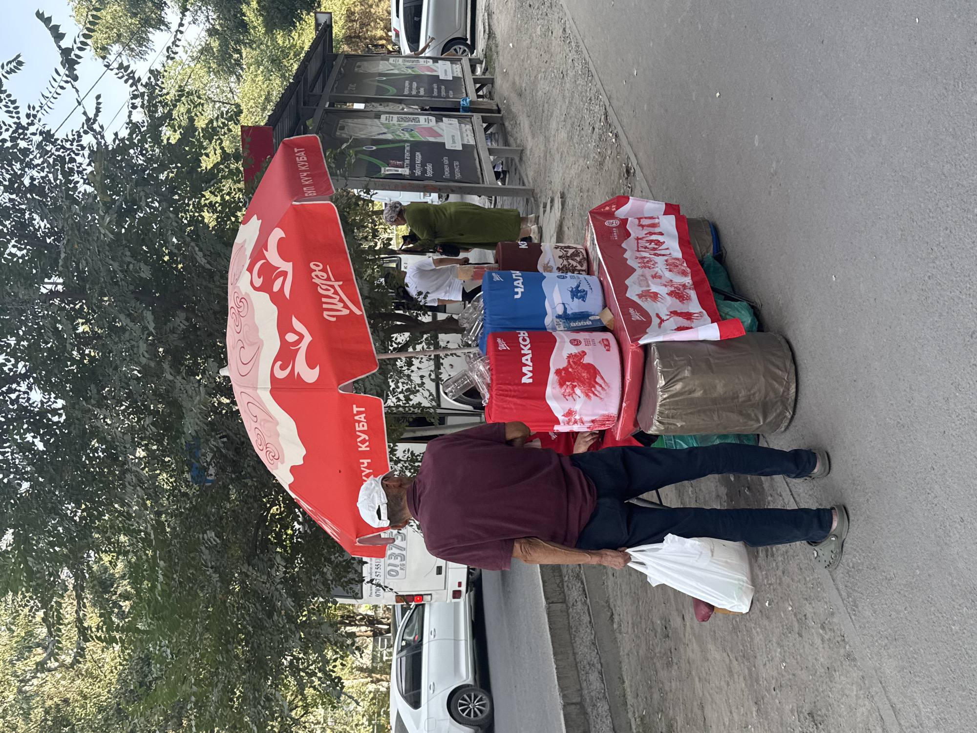 An elderly man wearing a maroon polo shirt, dark pants, and a white baseball cap stands facing a red umbrella with white lettering, holding a white plastic bag in his left hand. He is on a sidewalk next to a street in Bishkek, Kyrgyzstan. Behind him, a red and white umbrella is set up over a cart displaying three large blue and red bags with white lettering and images. The cart also has a red and white cloth draped over the front, depicting various scenes. To the right of the umbrella is a bus stop shelter with advertisements. Several cars are visible on the street, and trees line the sidewalk. The overall scene suggests an outdoor vendor or small market setup.