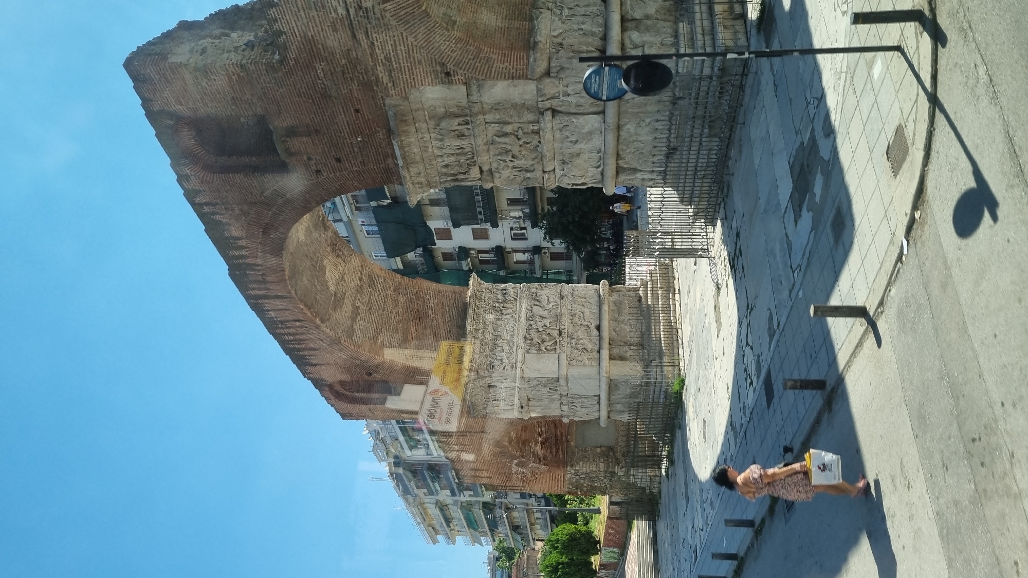 The photograph shows the Arch of Galerius in Thessaloniki, Greece, on a sunny day. A woman in a patterned dress and pink sandals is walking on the sidewalk to the left of the arch, carrying a white shopping bag. Behind the woman and the arch are multi-story buildings, some with green awnings. A street is in the foreground, with a shadow cast from a street lamp. The arch itself is made of brick and stone, with intricate carvings on the lower sections. The sky is clear blue.