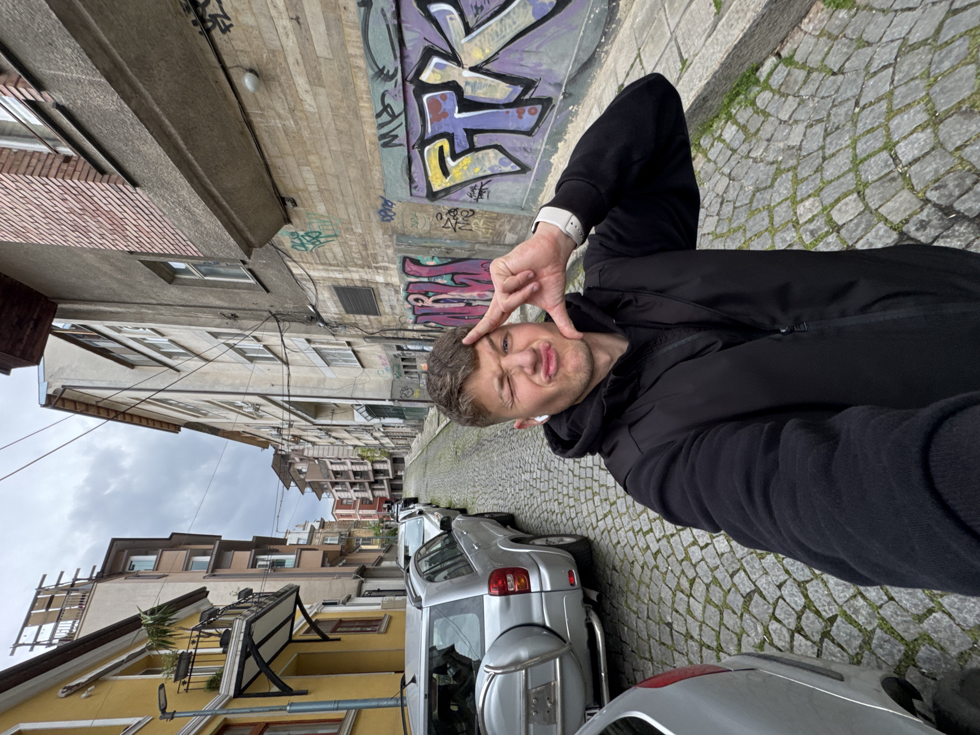 The image features a young man capturing a selfie on a cobblestone street in what appears to be Plovdiv, Bulgaria, on an overcast day. The man, positioned in the right foreground, is wearing a black hooded jacket and a white watch on his left wrist. He is making a playful or humorous facial expression with puckered lips and squinted eyes, and holds his left hand up near his face, forming a "V" or peace sign with two fingers.
Behind the man, a textured wall is adorned with vibrant, multi-colored graffiti. The stylized lettering includes discernible elements resembling 'T' and 'E' within the tags. The street is paved with characteristic cobblestones, with small tufts of grass visible between the stones.
The narrow street is lined with parked vehicles, including a silver SUV with a rear-mounted spare tire prominent on the left side. Tall, multi-story buildings flank the street, showcasing varied architectural styles; some sections feature exposed brick, while others are painted in light hues, including yellow, with visible windows and balconies. The sky above is cloudy, indicating an overcast day. The overall impression is a casual, street-level scene with an urban backdrop.